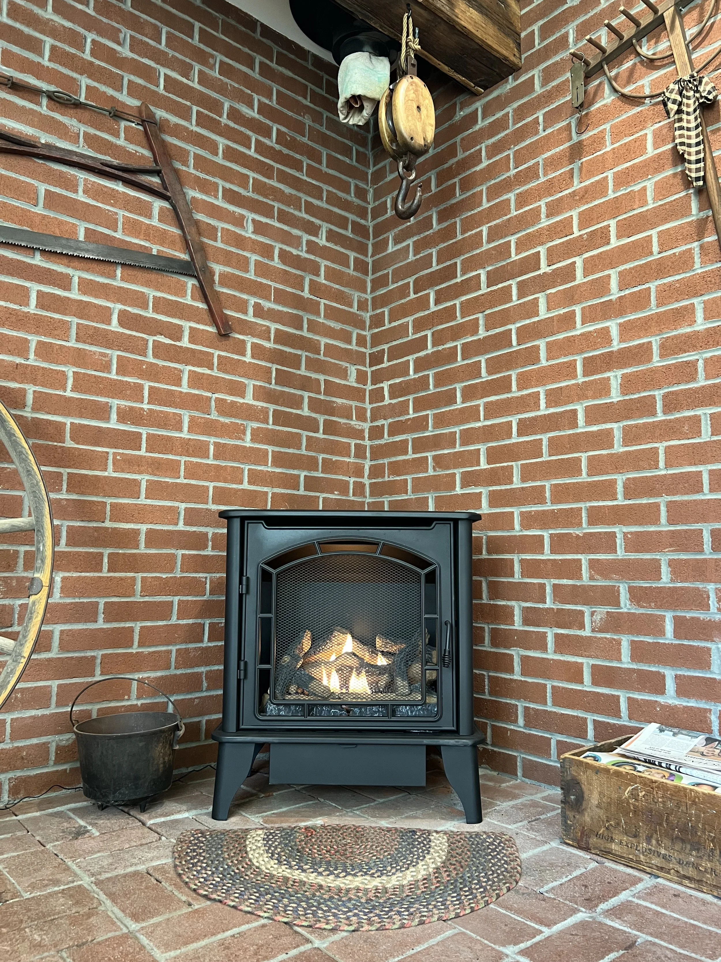 A cozy corner with a brick fireplace and a fire burning inside. Above the fireplace, there are vintage tools and a pulley hanging on the wall. To the left, part of a rusty saw is visible. On the floor, in front of the fireplace, is a round braided rug. To the right, there is a wooden box filled with newspapers. A small, black bucket is placed on the floor near the wall.
