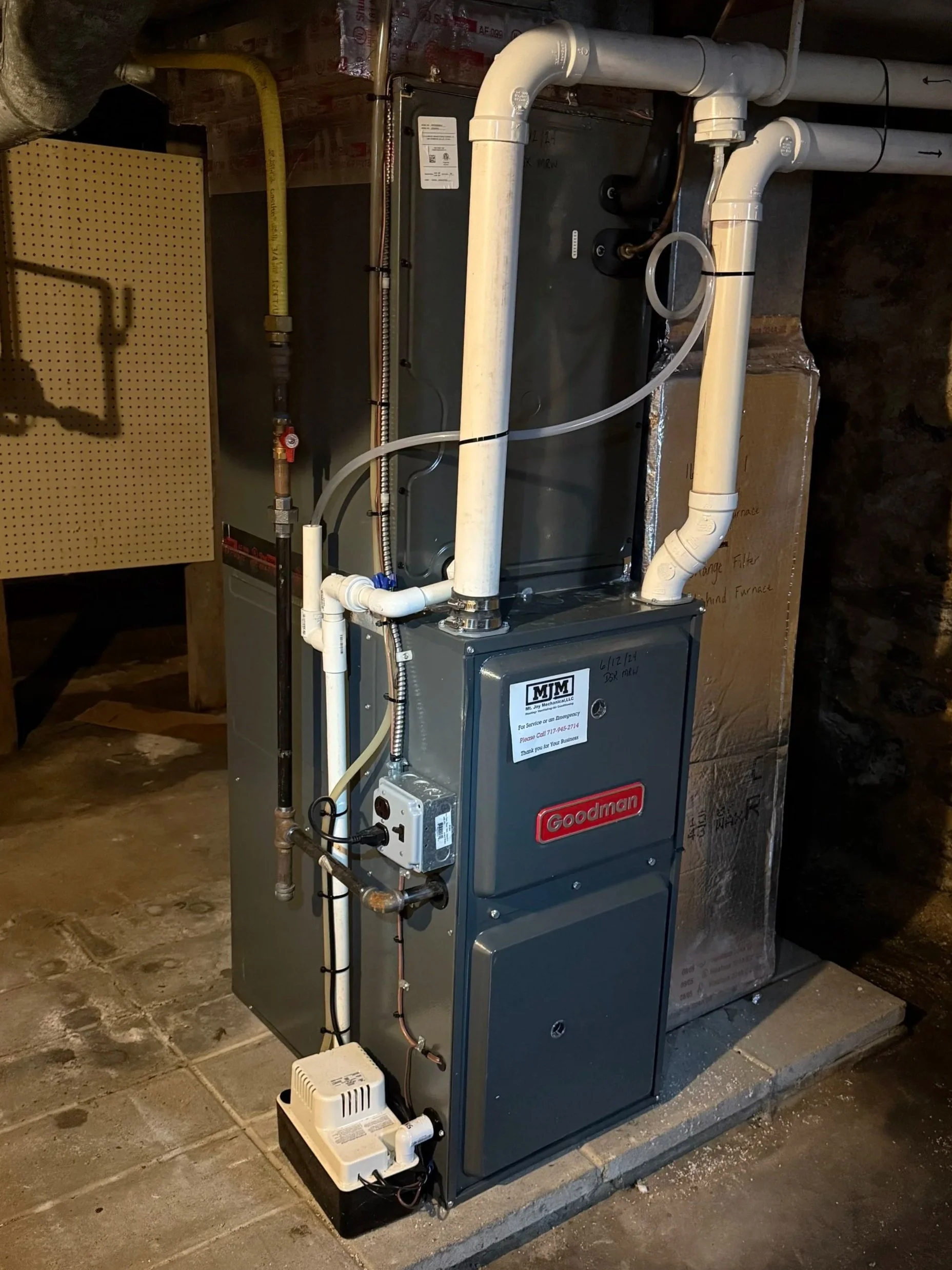 A Goodman HVAC furnace unit with connected pipes, electrical wiring, and control components installed on a concrete floor in a basement or utility room.
