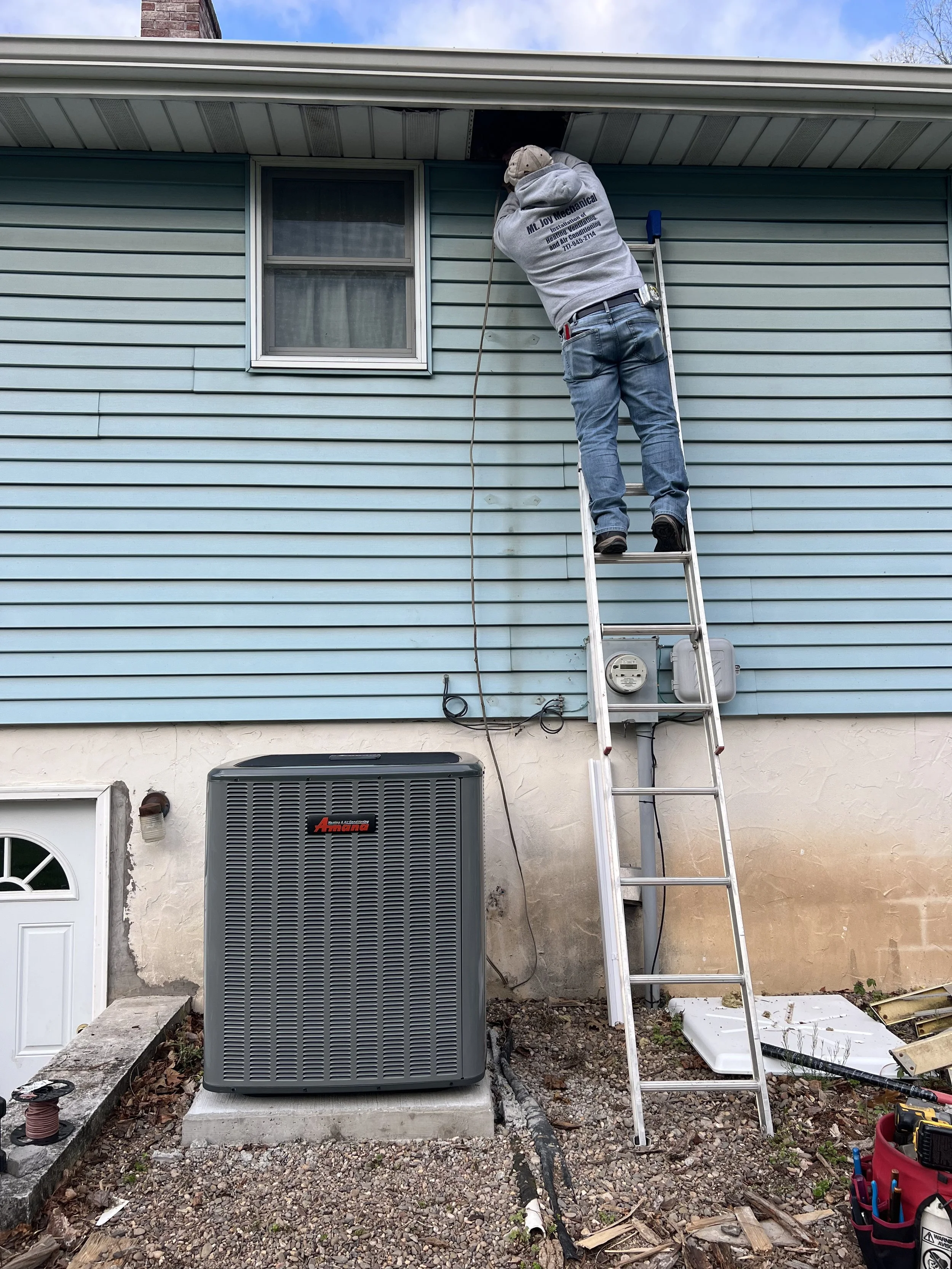 A worker on a ladder installing or repairing an outdoor electrical or utility box on the side of a house with blue siding.