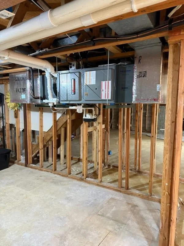 Interior of a house under construction showing exposed wooden framework, HVAC ductwork, and plumbing piping.