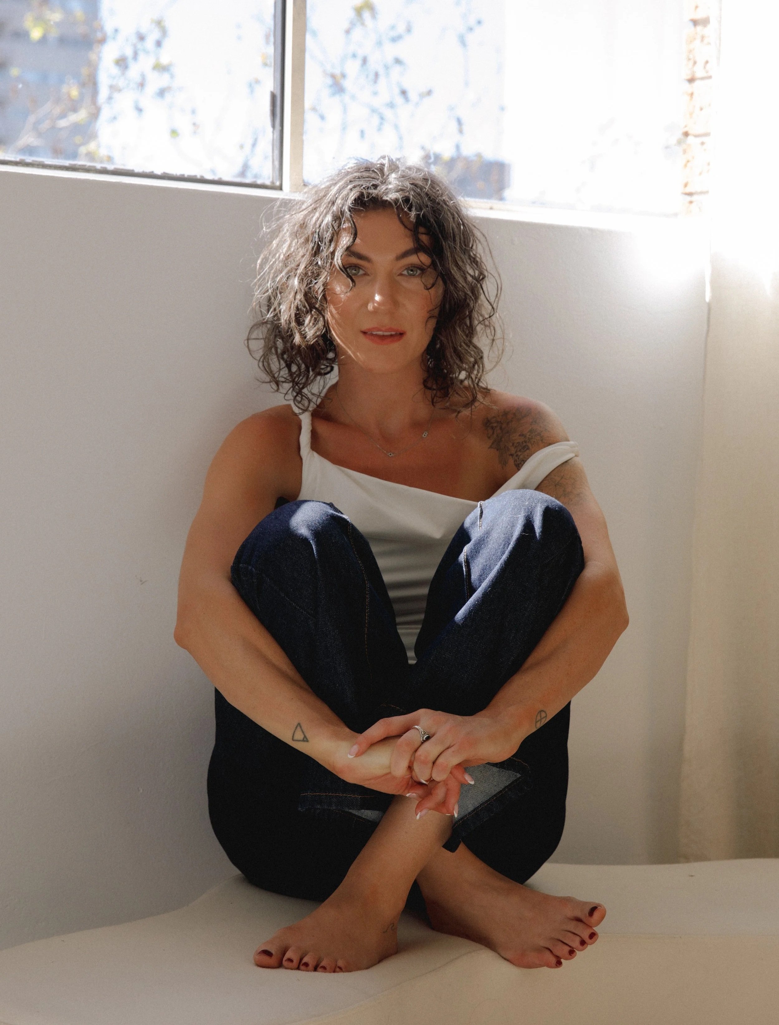 A woman with curly hair sitting on the floor by a light-colored wall and window, wearing a white tank top and dark jeans, with tattoos on her arms and foot, looking at the camera.