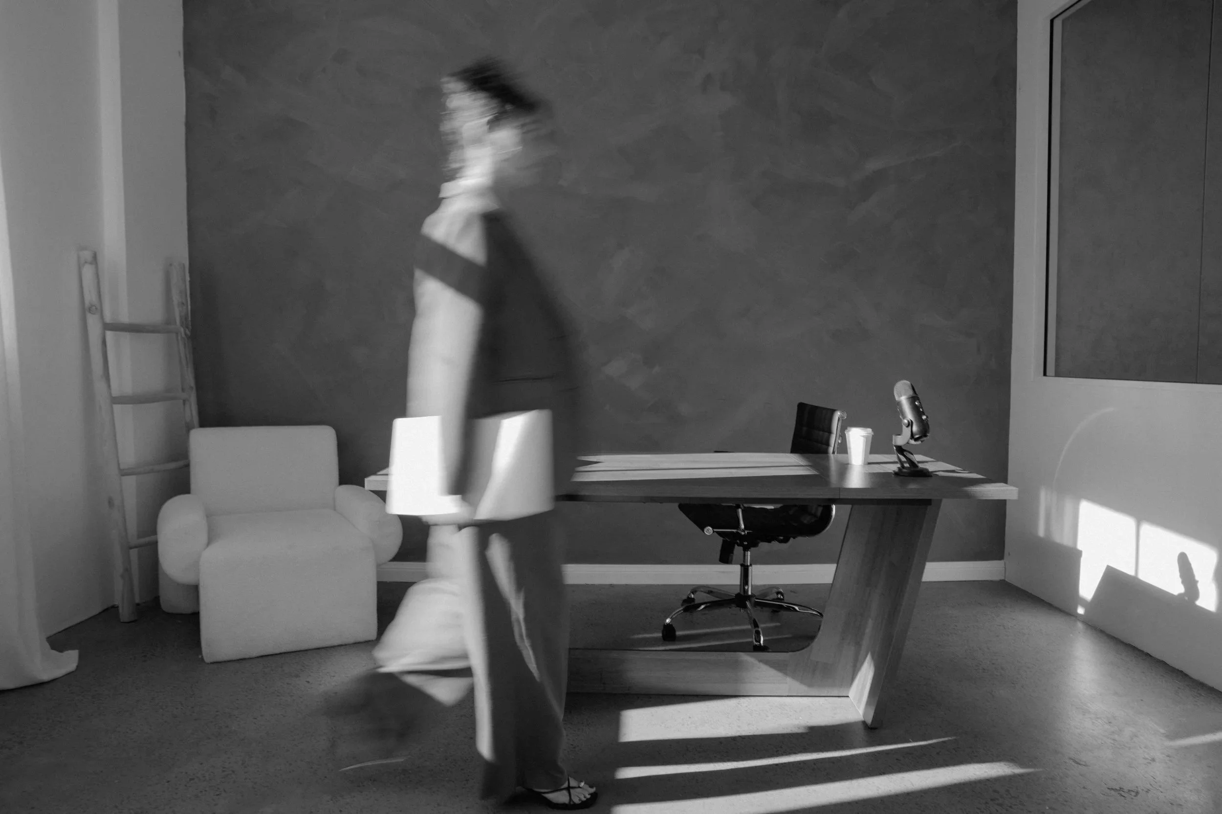 A woman in a dress and sandals is walking through an office space with a desk, a chair, a microphone, and a cup on the desk. The office has a textured wall, a ladder, and a white armchair.