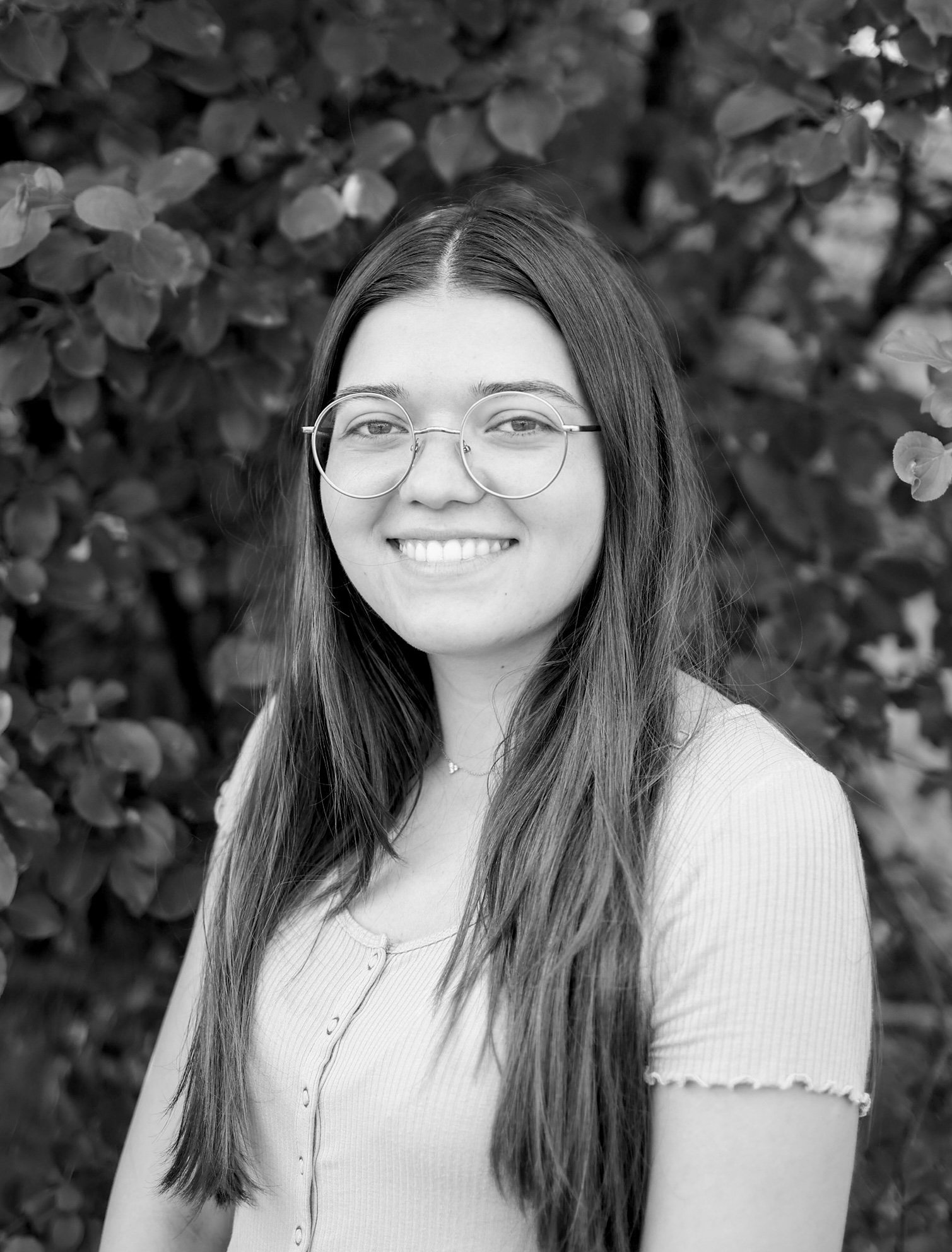 Black and white portrait of a young woman with long hair and glasses, smiling in front of leafy background.