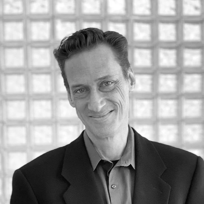 A black-and-white portrait of a smiling man dressed in a suit jacket and collared shirt, standing in front of a modern glass block wall.
