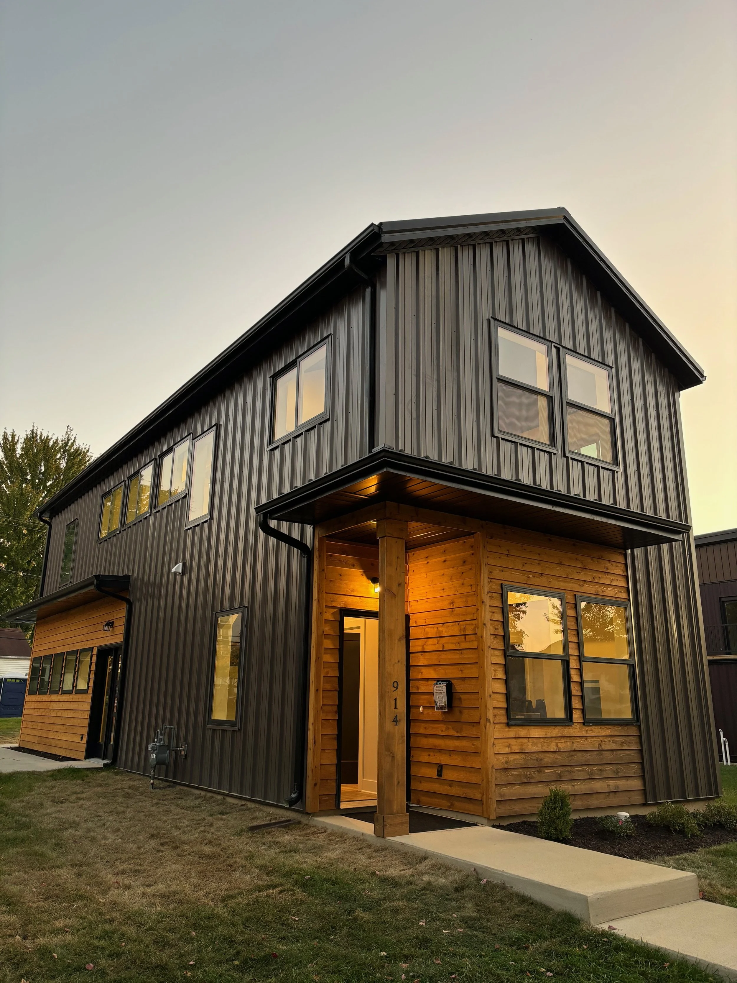 Modern multi-story house with black metal siding and wood accents, illuminated porch, and multiple windows