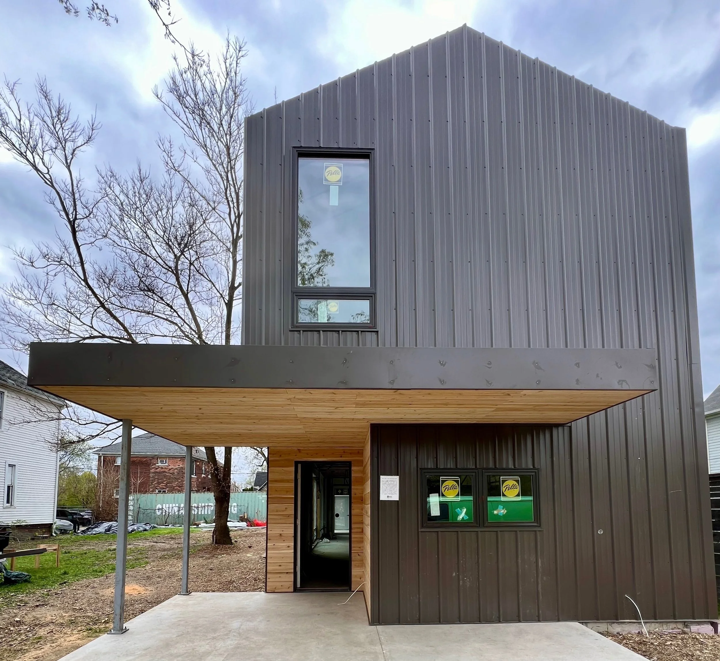 A modern, two-story building with dark gray and wood exterior paneling, featuring large windows, with a covered entrance area supported by metal posts, and a construction site surrounding it.