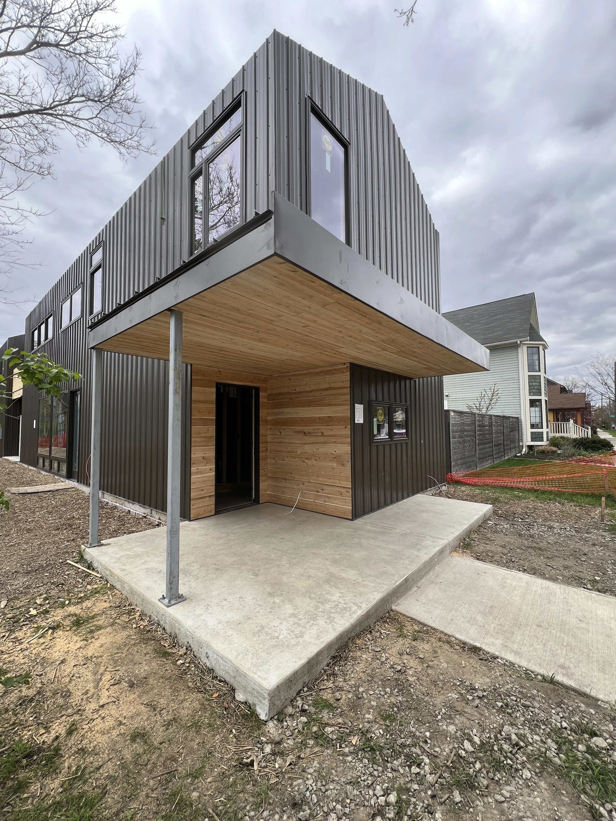 Modern two-story house with black metal exterior, wooden accents, and a small covered porch area under an overhanging second floor, set in a residential neighborhood with partly cloudy sky.