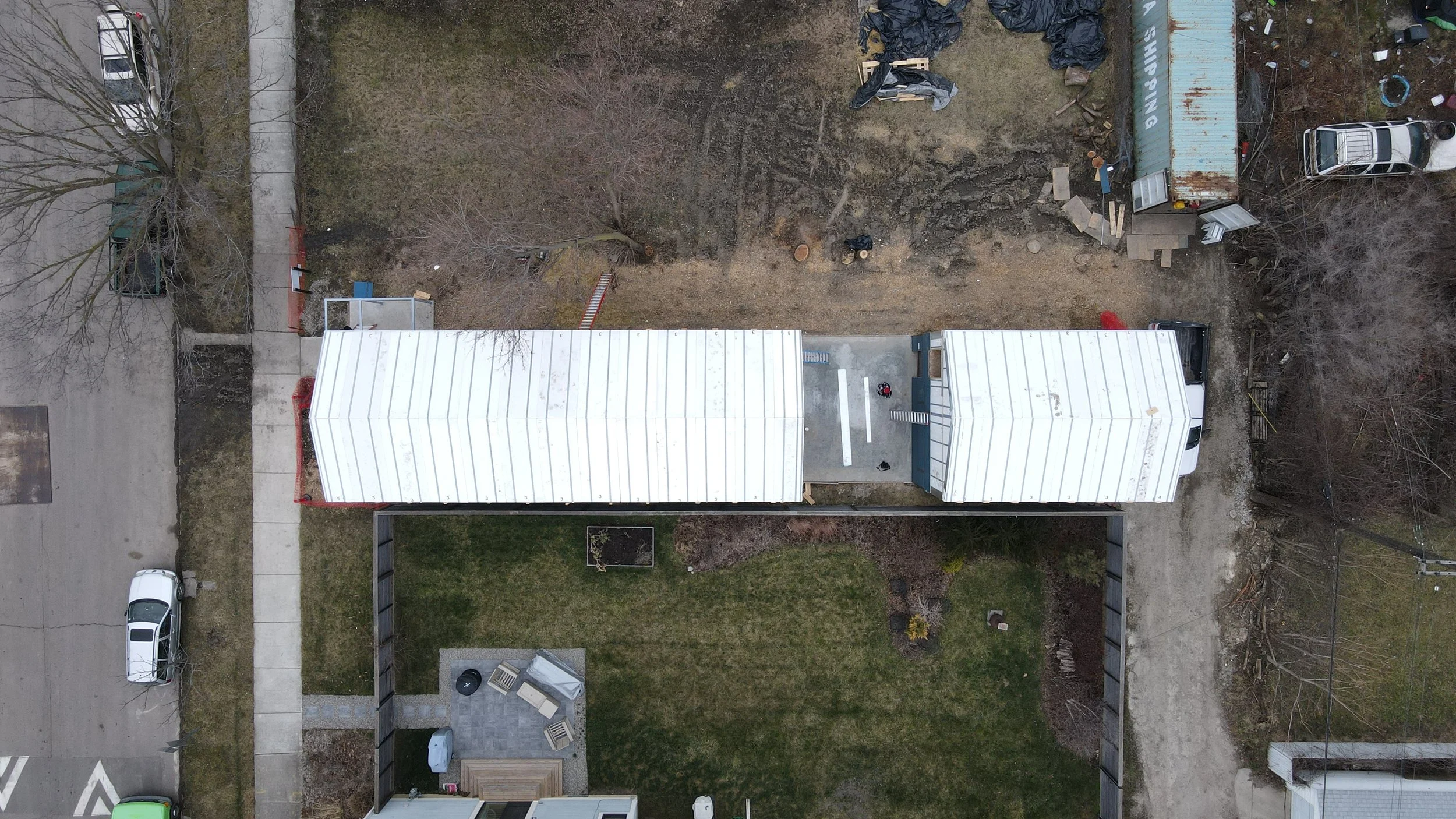 An aerial view of a backyard with a new white metal roof structure, a green lawn, a patio with furniture and a grill, and construction area with dirt, materials, and equipment.