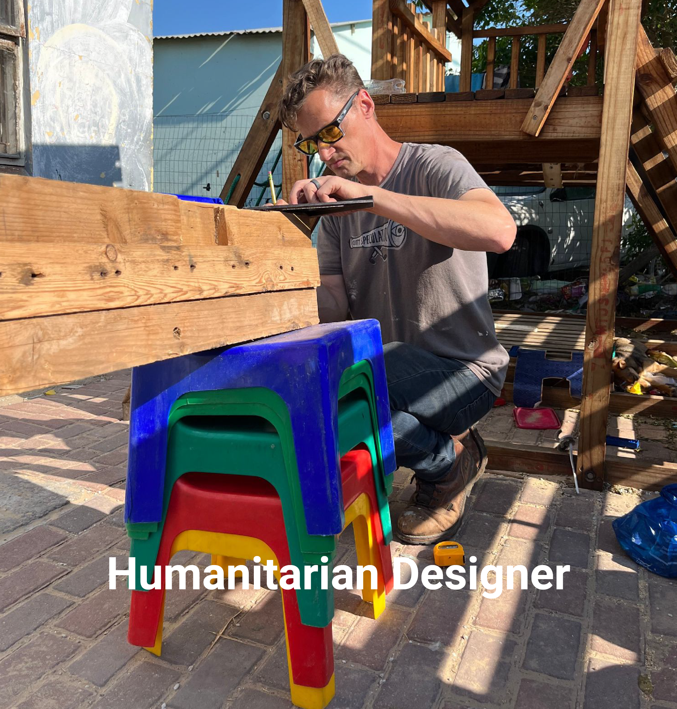 A man working on a woodworking project outdoors, surrounded by colorful plastic chairs, with a wooden structure under construction nearby.