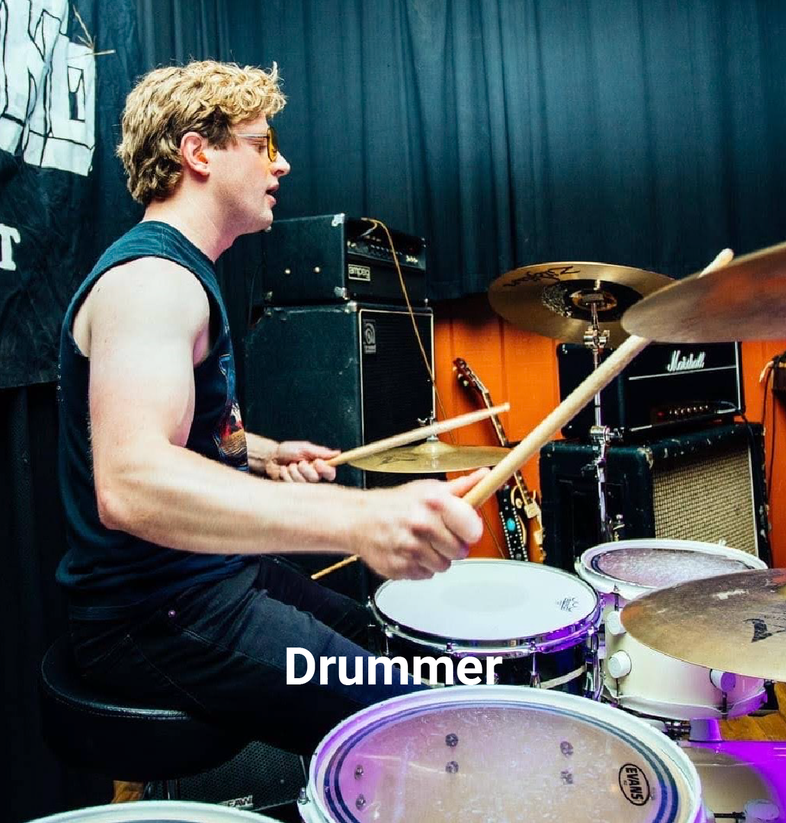 A young man playing drums in a music studio, wearing sunglasses and a sleeveless shirt, with amplifiers, guitars, and black curtains in the background.