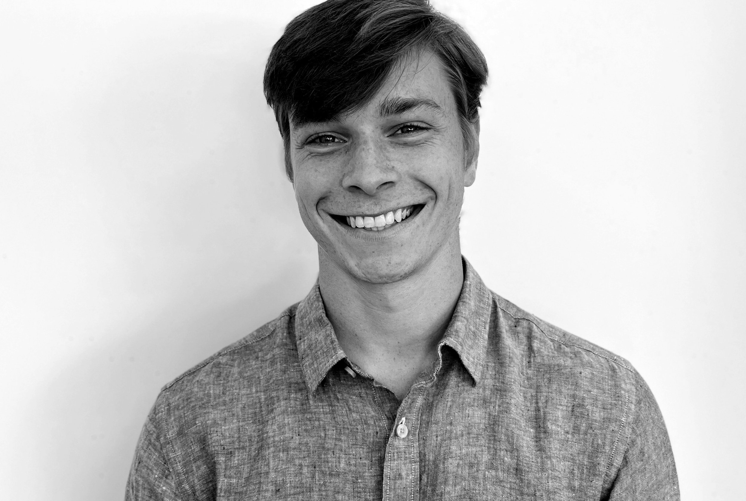 Black and white photo of a young man with dark hair smiling and wearing a button-up shirt, standing against a plain background.