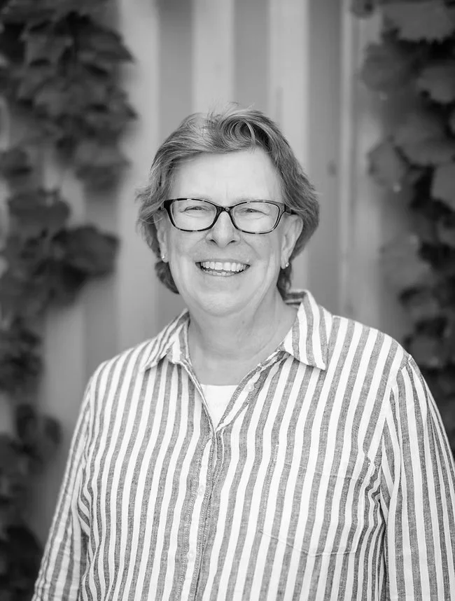 A smiling woman with short hair and glasses wearing a striped button-up shirt, standing outdoors with blurred leafy background.