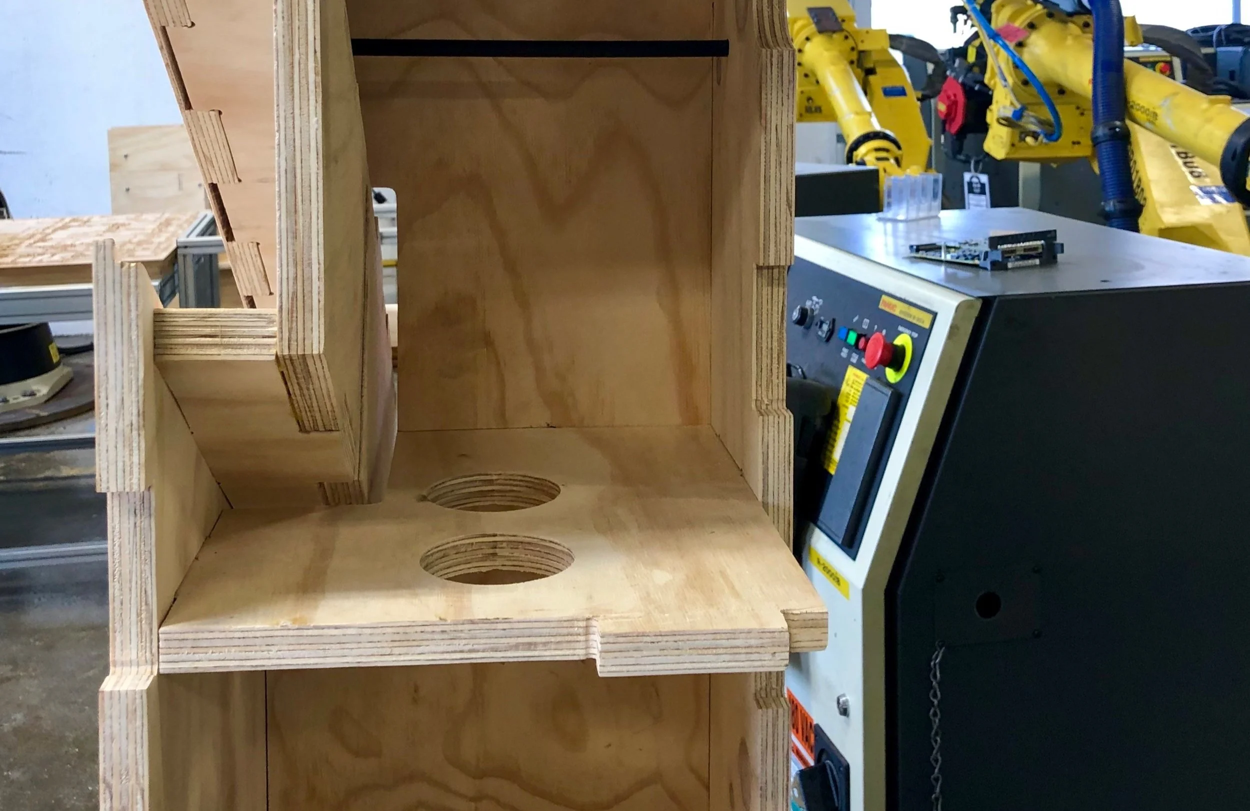 Unfinished wooden birdhouse with two circular entry holes on the base, in a workshop with tools and machinery in the background.