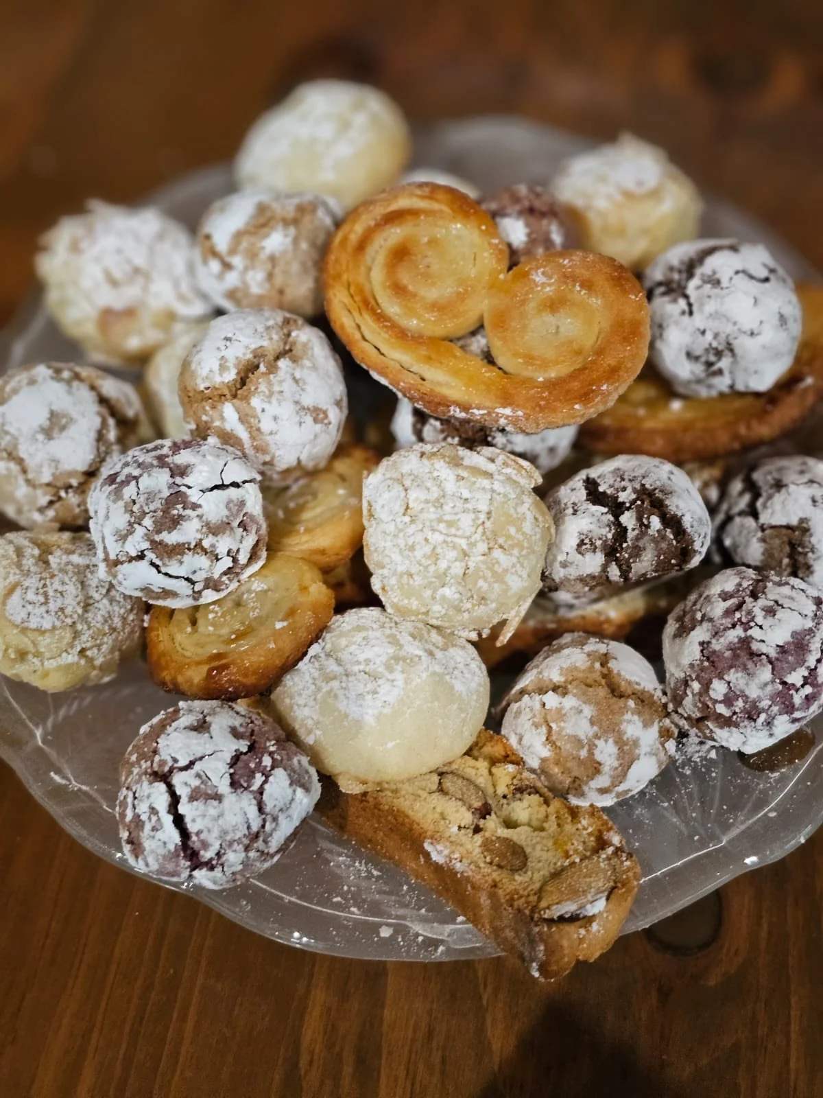 Assorted cookies and pastries on a glass plate, including chocolate crinkle cookies, cream-filled sandwich cookies, and snickerdoodles, dusted with powdered sugar.