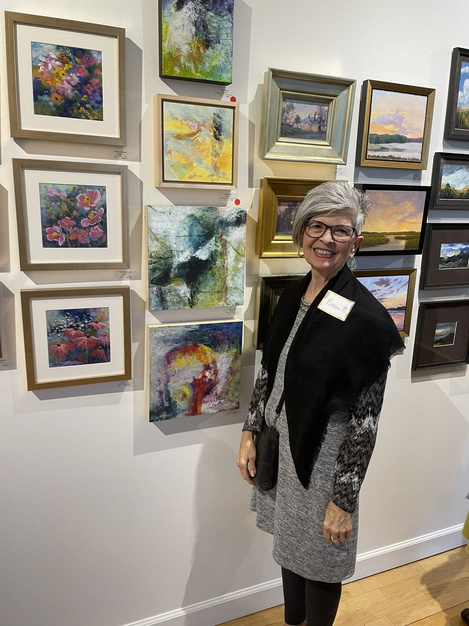 An older woman with short gray hair and glasses smiling at an art gallery, standing in front of a wall displaying various colorful paintings and framed artwork.