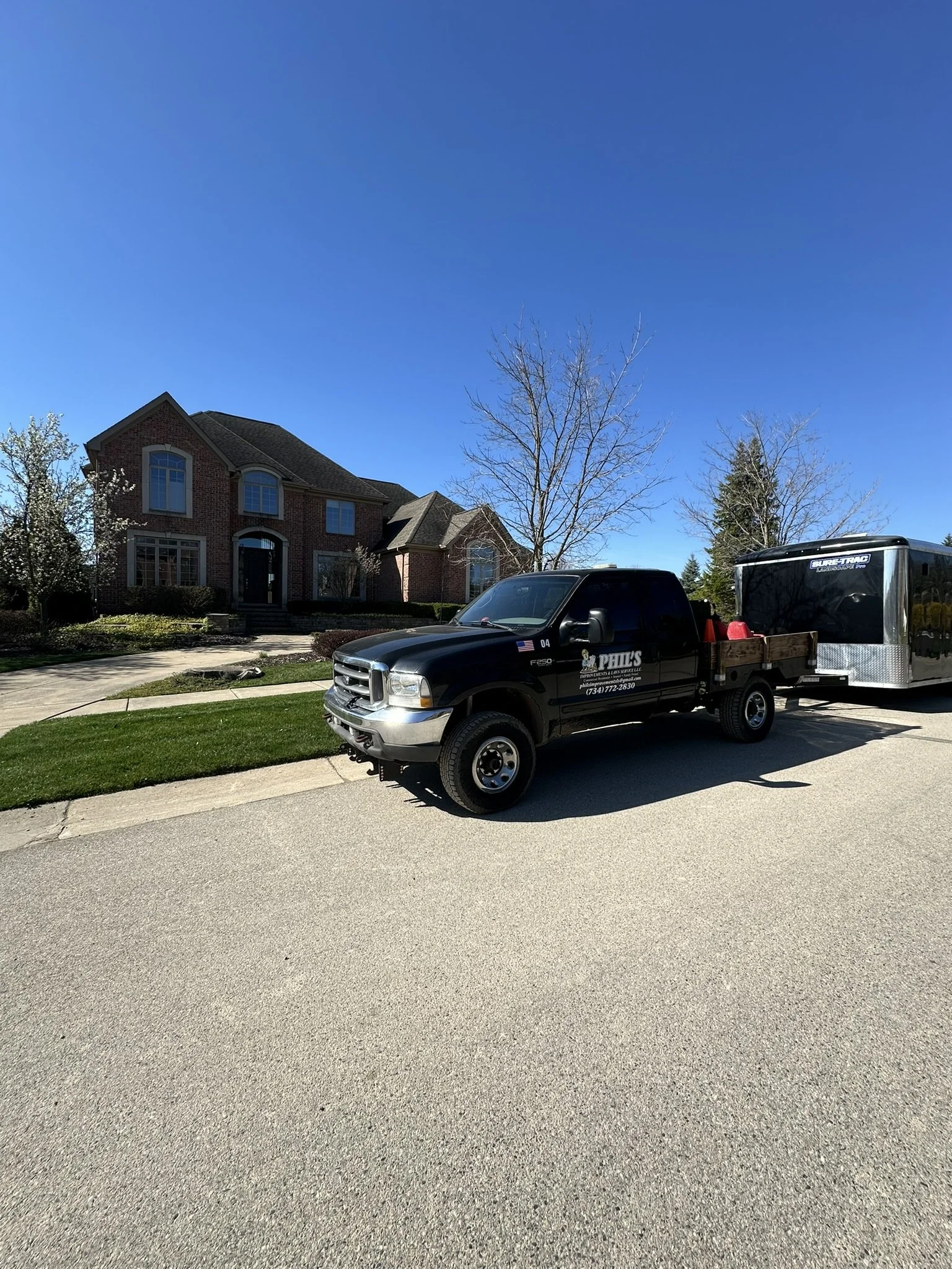 A black pickup truck with a trailer attached parked on a residential street in front of a brick house with a front yard and a sidewalk, under a clear blue sky with leafless trees.