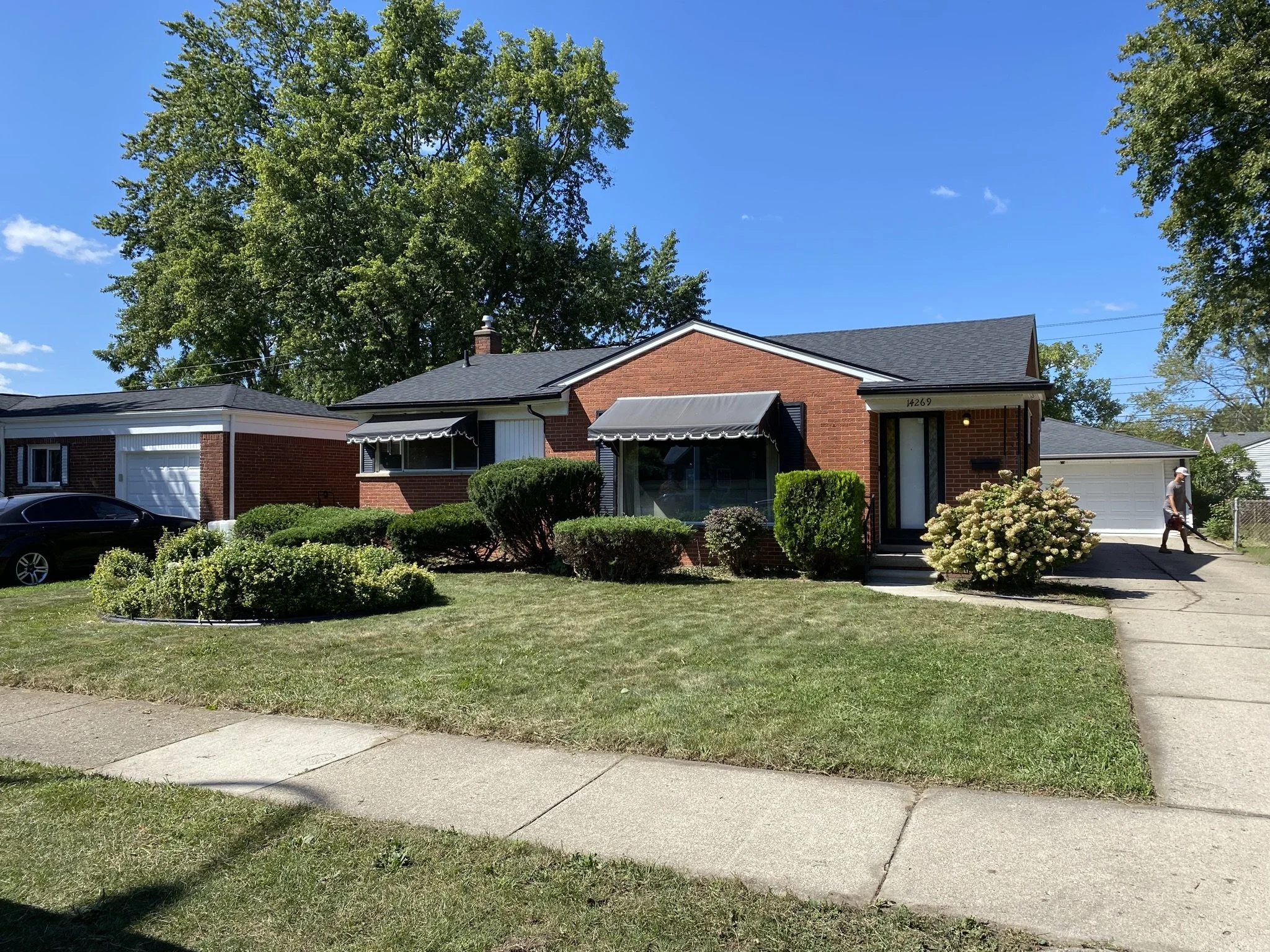 A single-story brick house with black awnings over the windows, surrounded by a well-maintained lawn and bushes, with a sidewalk and driveway, and a person walking with a shopping bag near the driveway, under a clear blue sky.