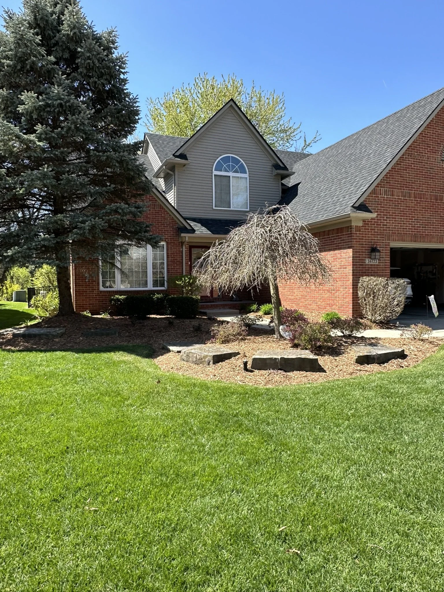 Front view of a brick and gray house with a landscaped yard, a large evergreen tree on the left, a smaller leafless tree in the front yard, and a clear blue sky.