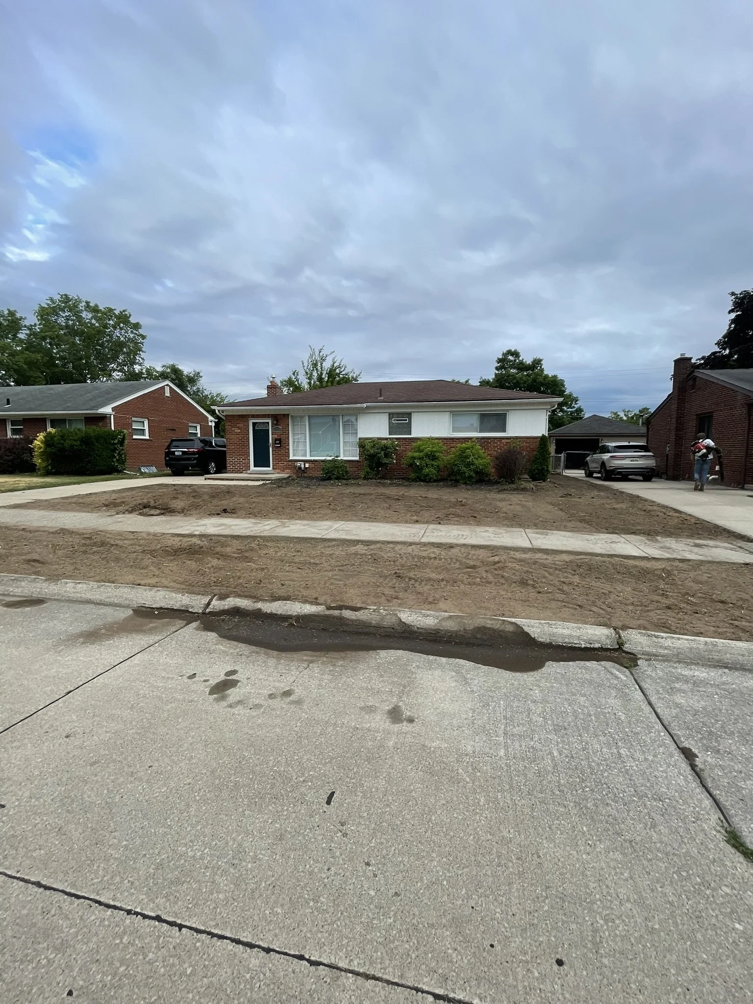 Front view of a house under construction with a dirt yard, sidewalk, and concrete driveway, overcast sky.