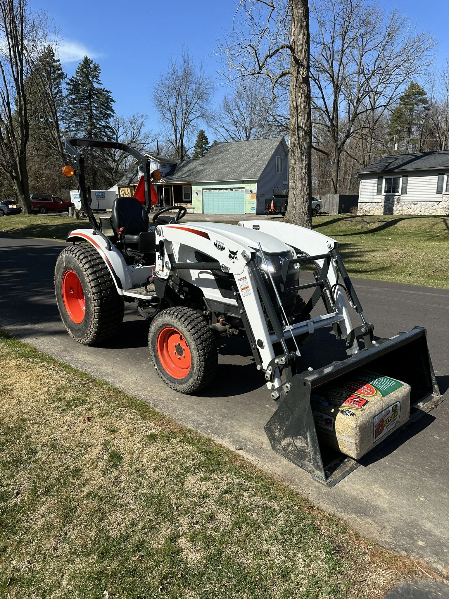 White compact utility tractor with a front loader attachment parked on a residential street with houses and trees in the background.