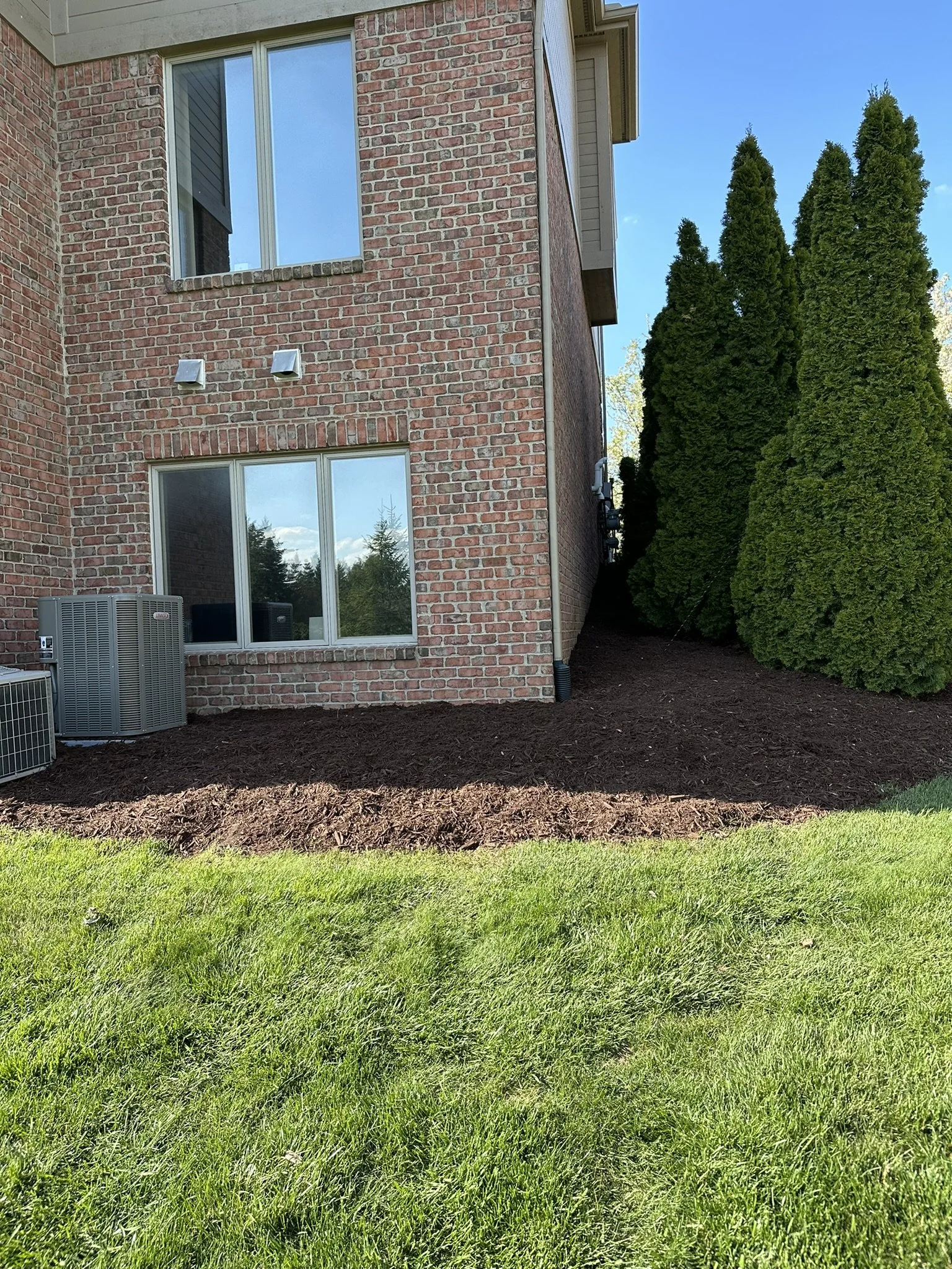 A side view of a brick house with three windows and an air conditioning unit. There are four large green trees on the right side, and a freshly mulched garden bed with green grass in the foreground.