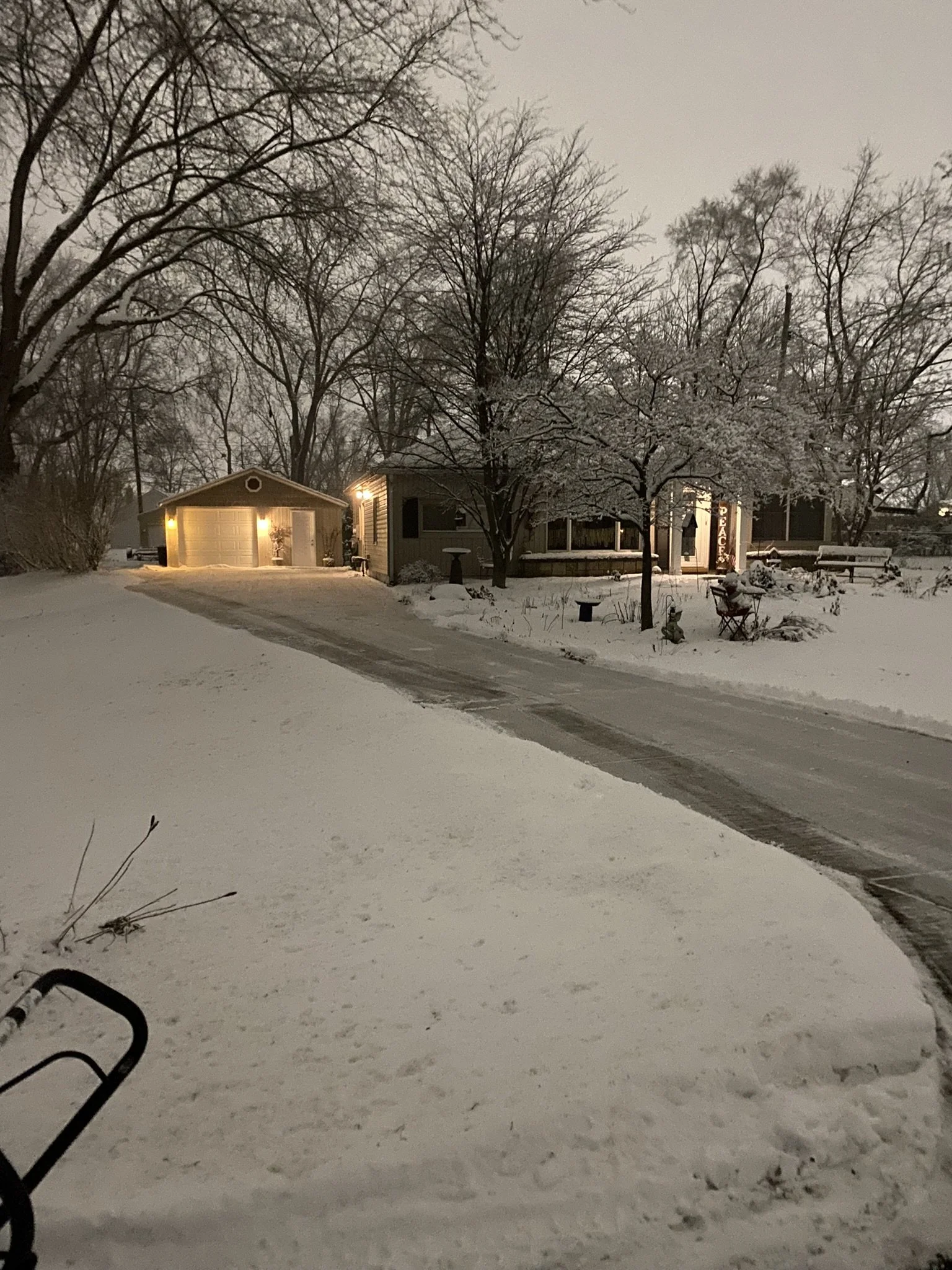 A snow-covered residential street at dusk with trees, houses, and a driveway leading to a garage with warm outdoor lights.
