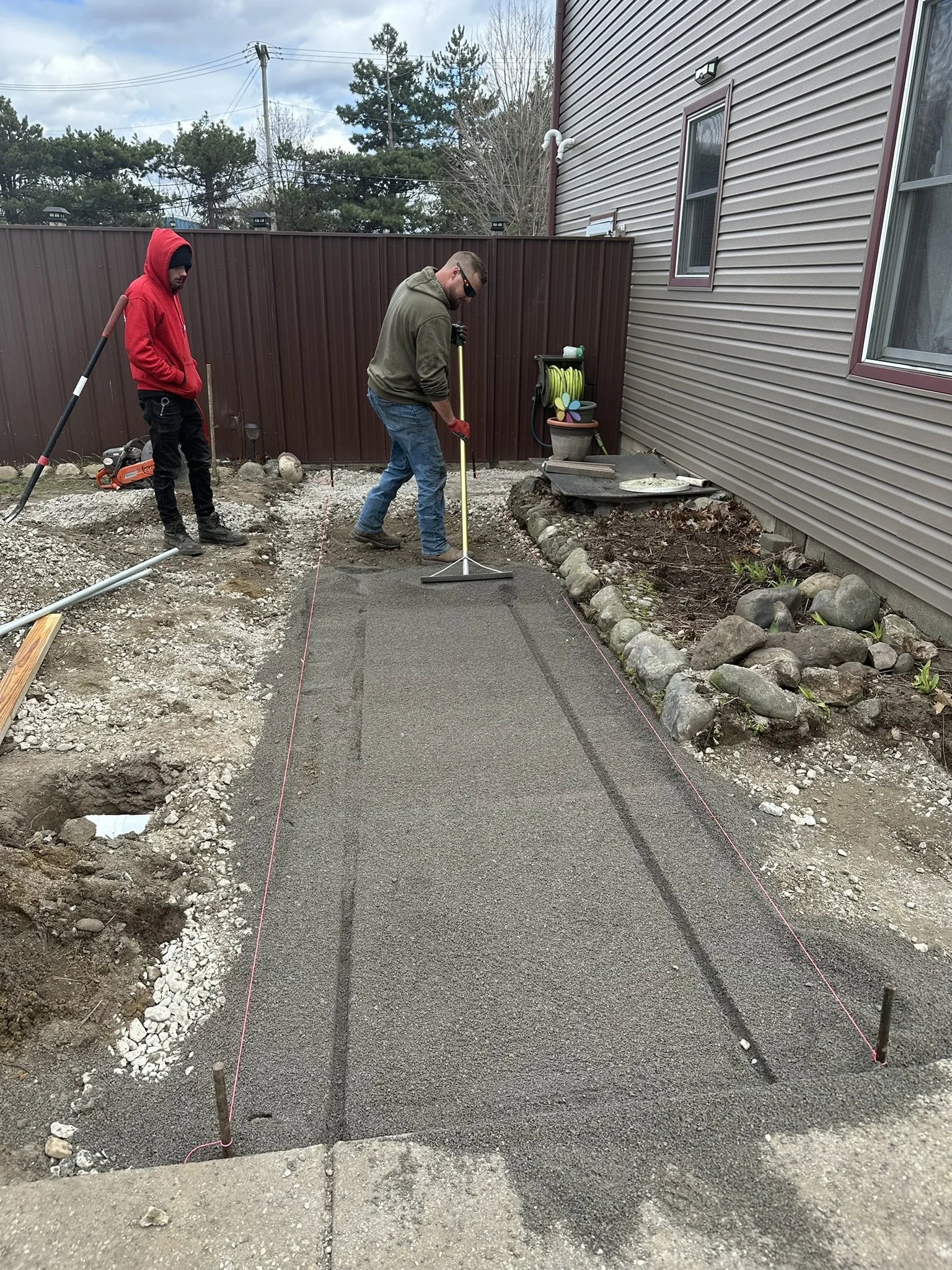 Two men are working on a small concrete sidewalk outside. One man is smoothing the wet concrete while the other stands nearby holding a tool. The area is marked with pink string lines for construction.