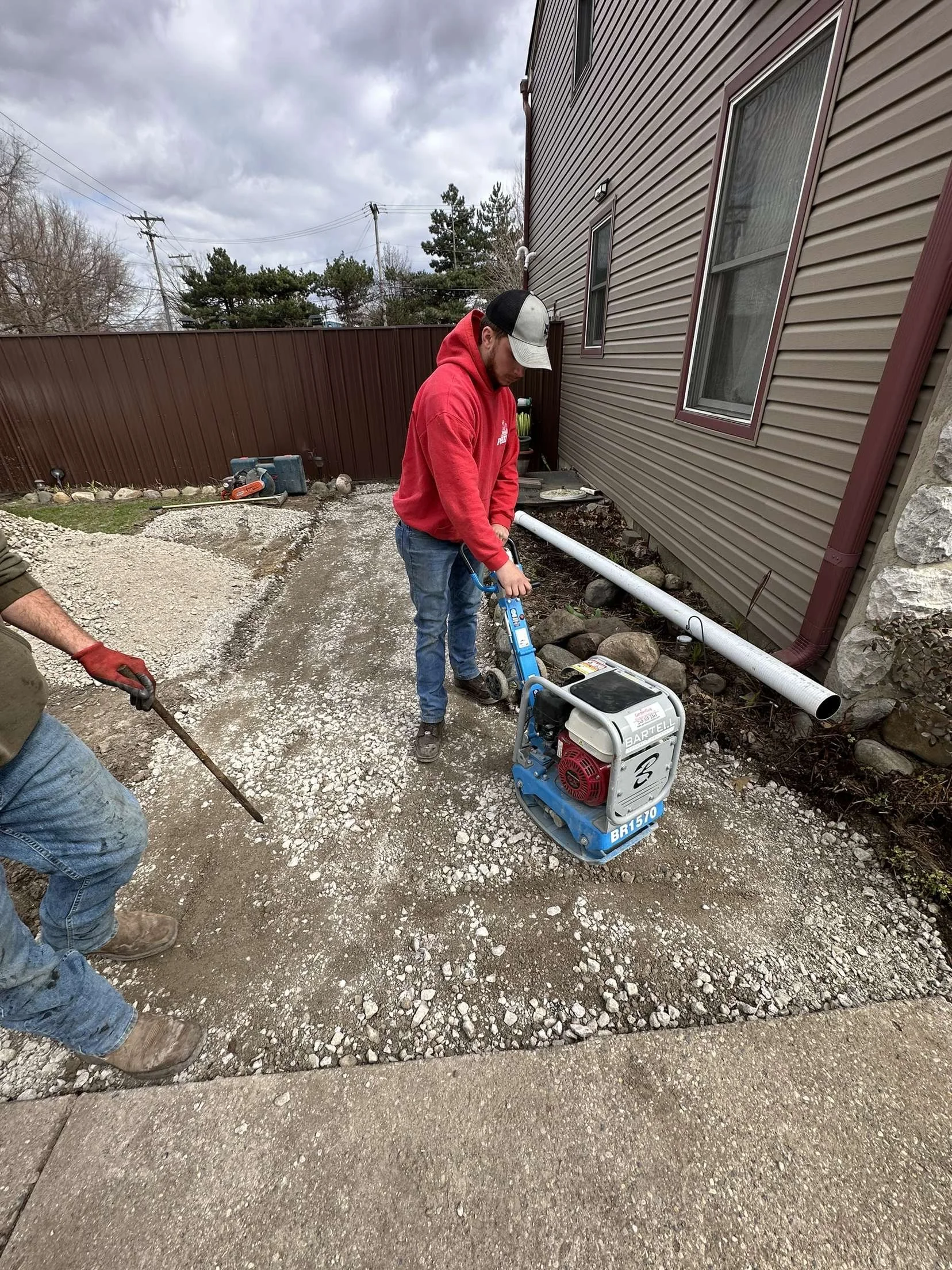 Two men working outdoors on a gravel pathway near a brown house, with one man operating a plate compactor and the other holding a tool, during cloudy weather.