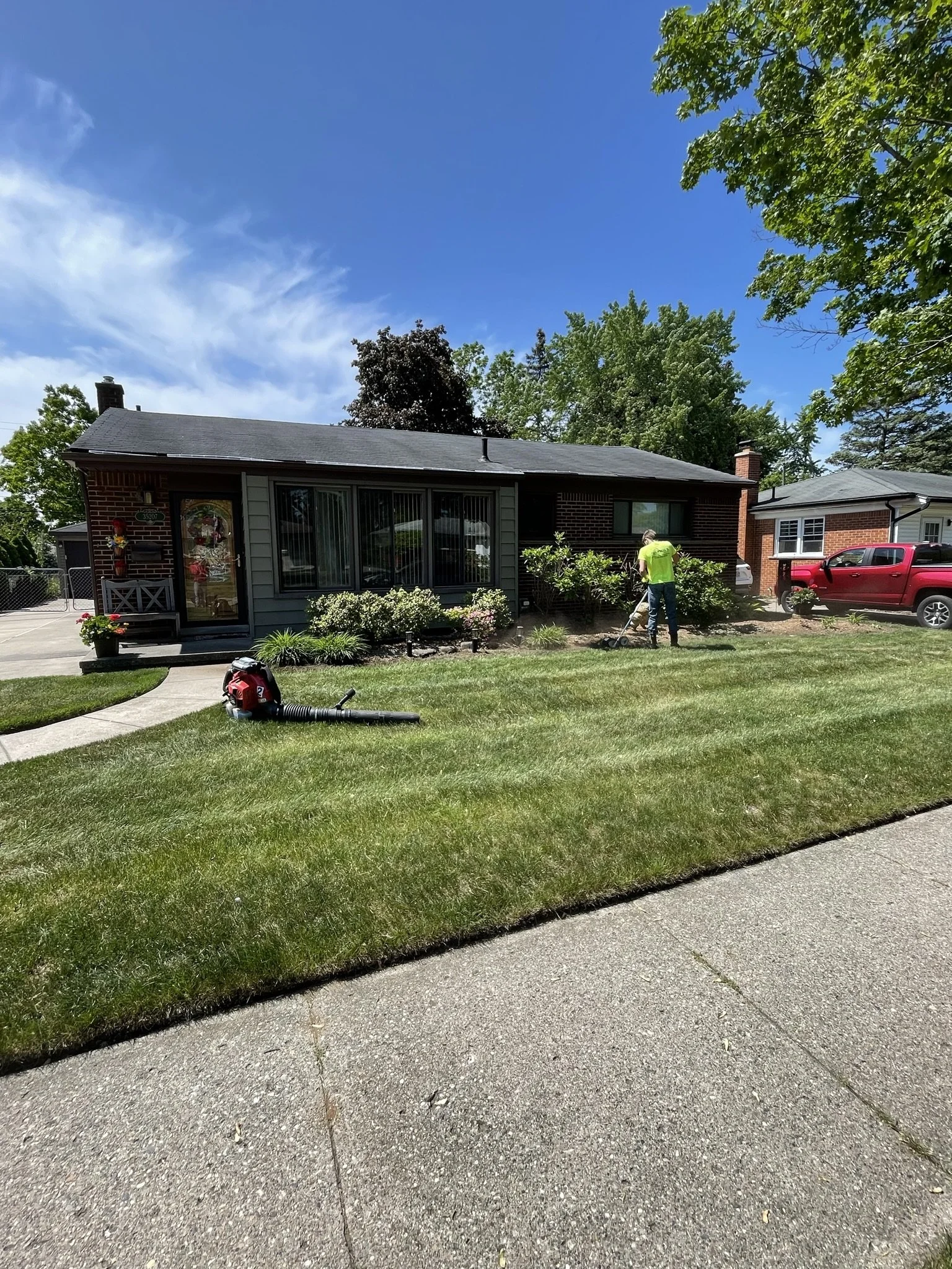 A man is trimming bushes in front of a single-story brick house with gray siding, surrounded by a green lawn and trees. A leaf blower is on the grass, and a red car is parked in the driveway.