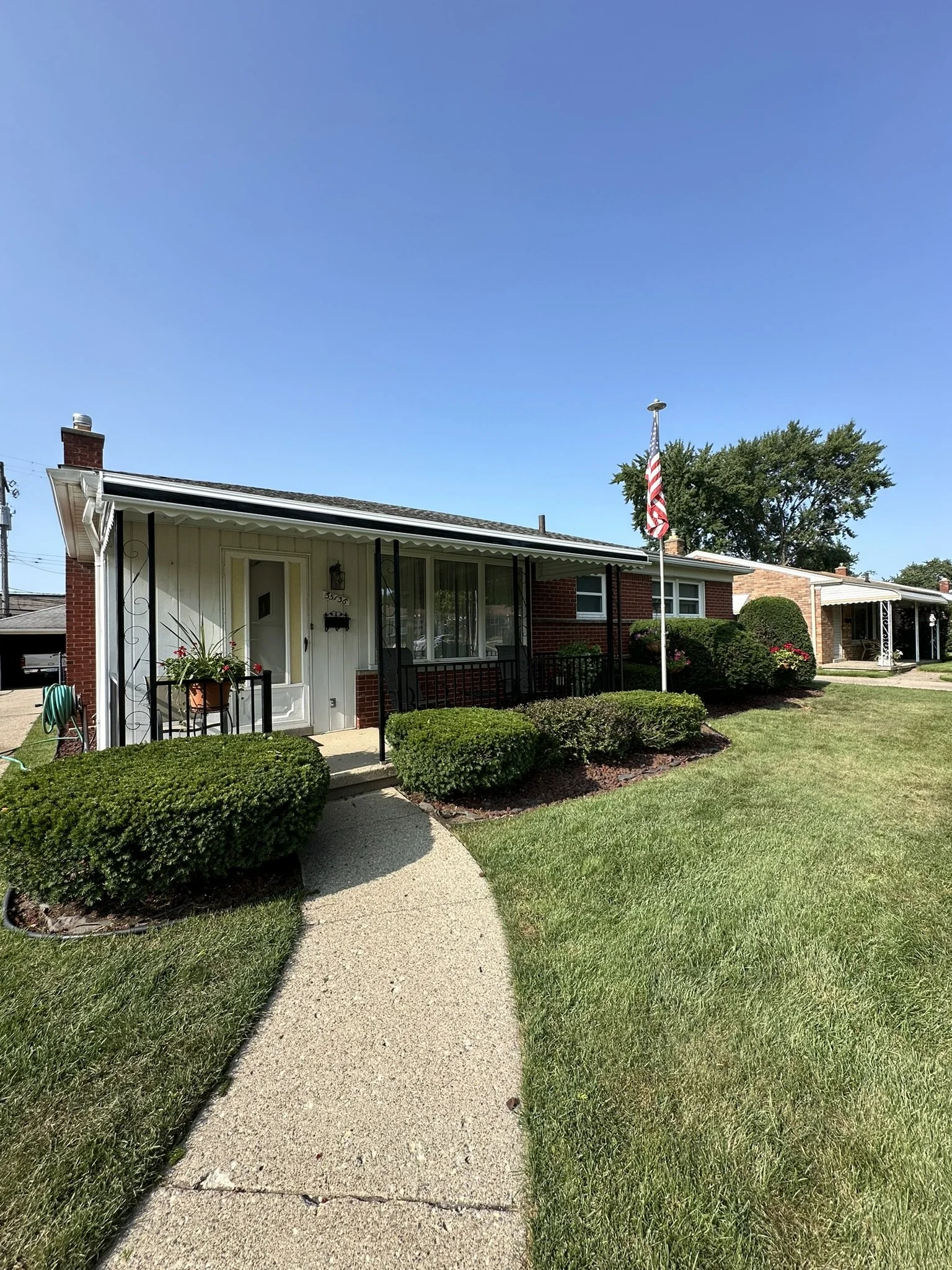 Front view of a single-story house with a curved concrete walkway, well-manicured lawn, bushes, a porch with two chairs, and an American flag on a flagpole.