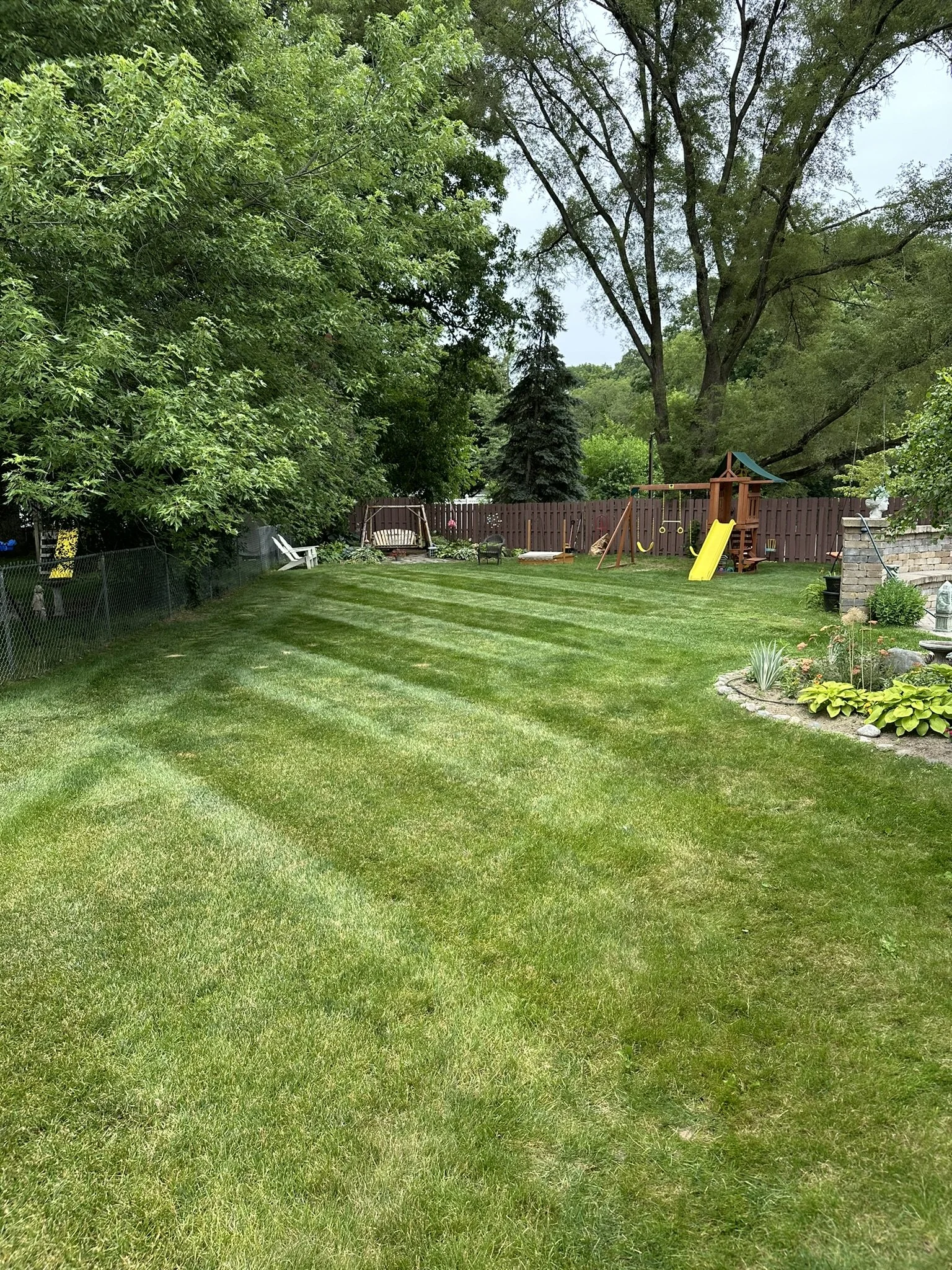A well-maintained backyard with lush green grass, surrounded by trees and a wooden fence. There is a children's playset with a slide, swings, and a sandbox, as well as garden beds with plants and flowers.
