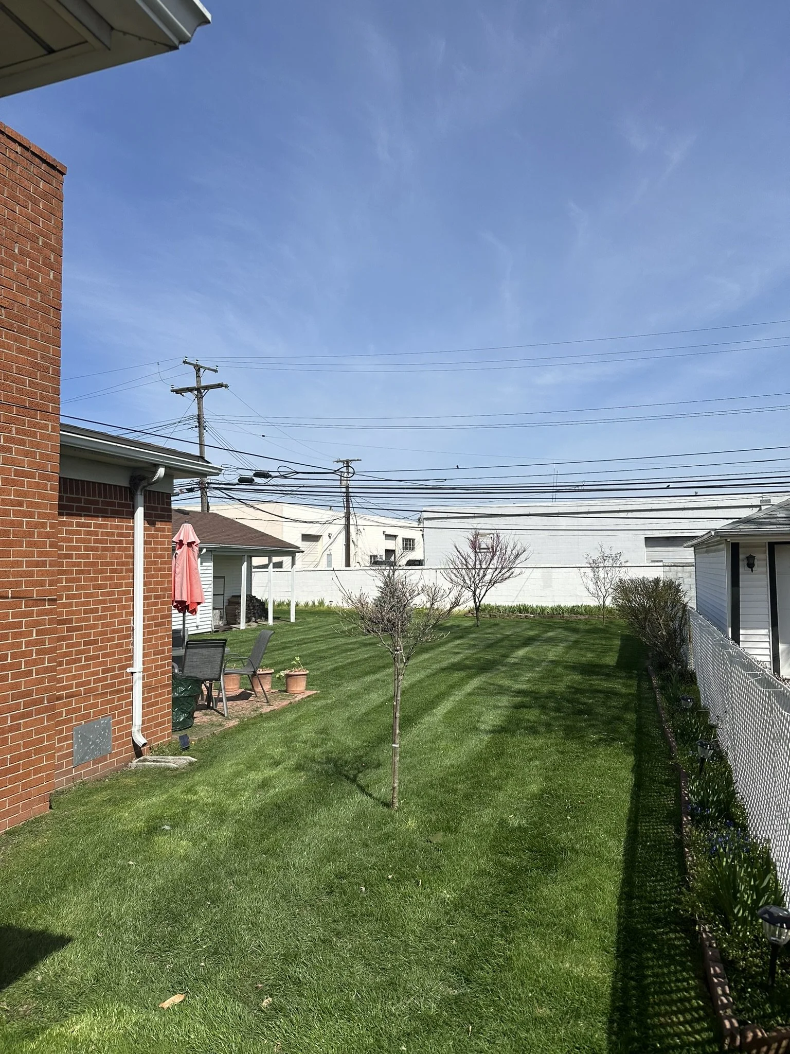 Backyard with a green lawn, small trees, patio furniture, a pink umbrella, and a white fence under a blue sky.