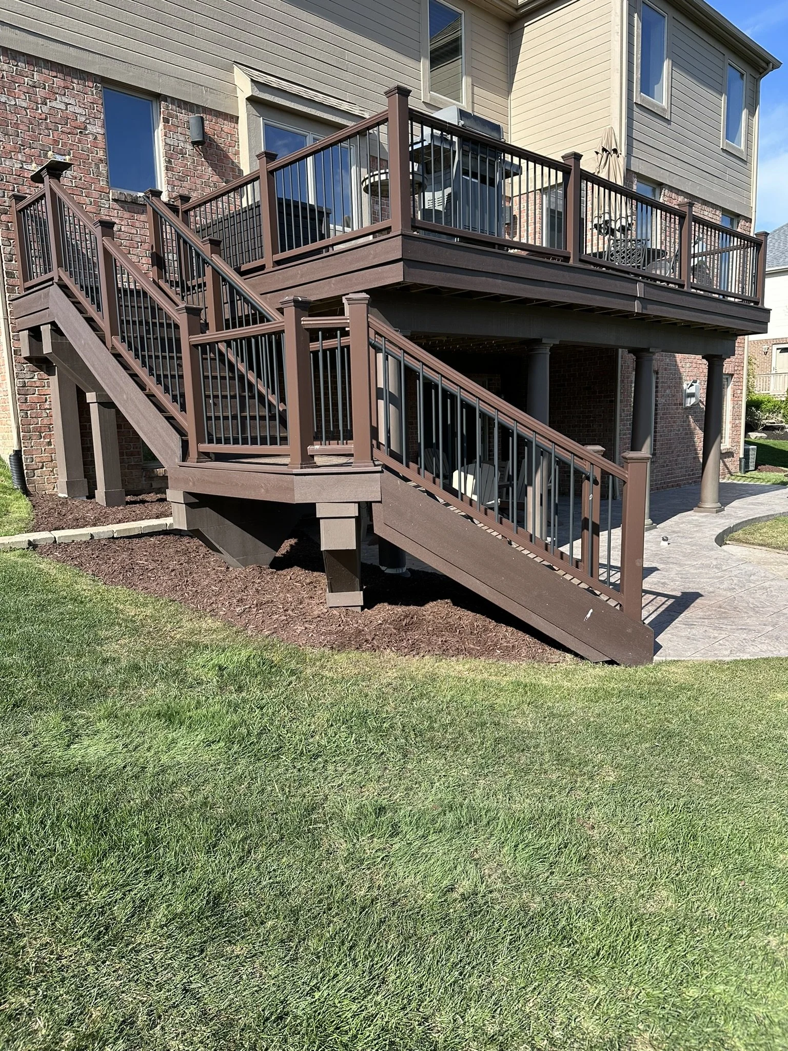 A multi-level wooden deck attached to the back of a house, with stairs leading down to a well-maintained lawn and a stone patio area.