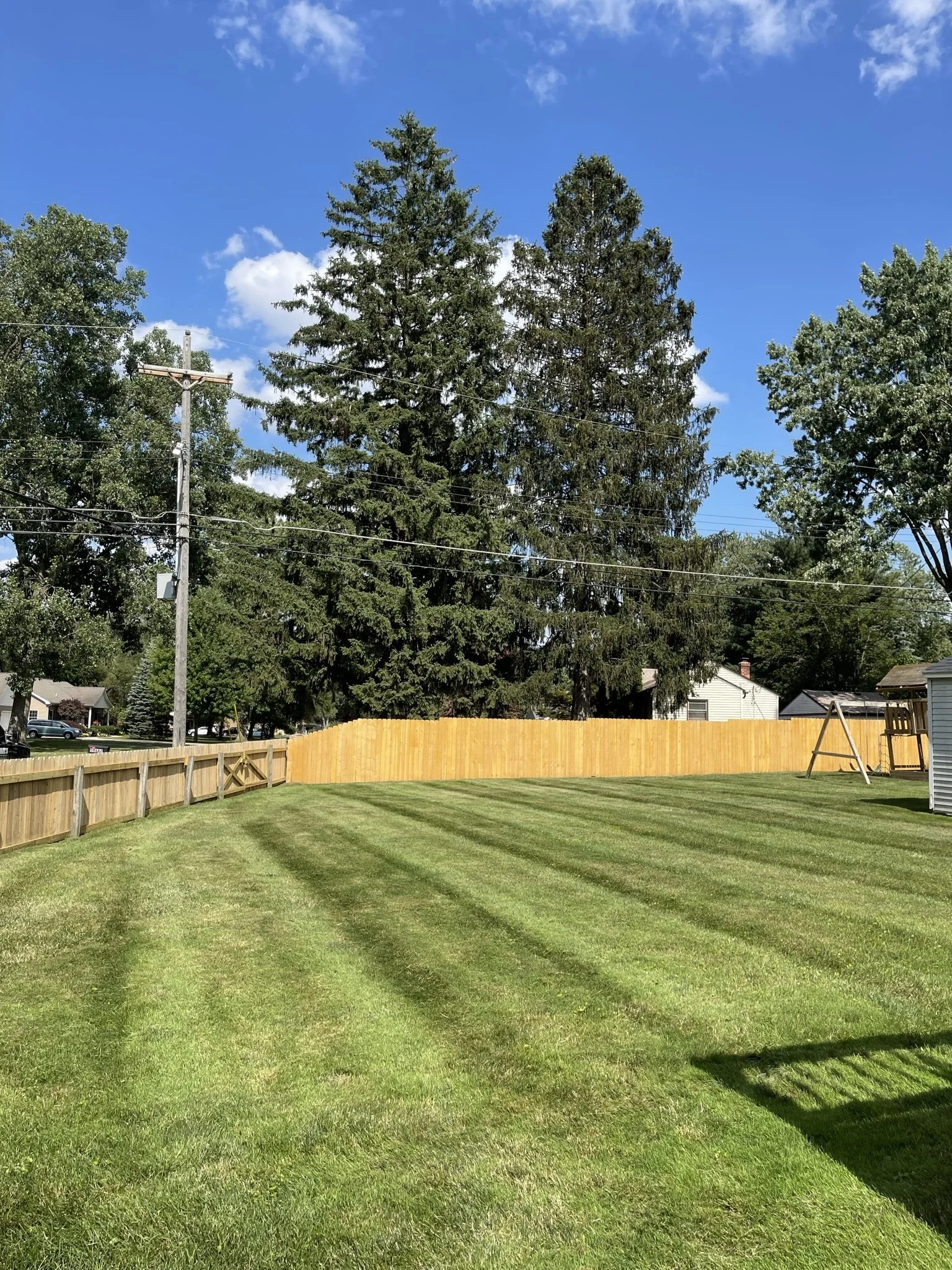 A backyard with a well-maintained grassy lawn, surrounded by a wooden fence and tall trees under a partly cloudy blue sky.