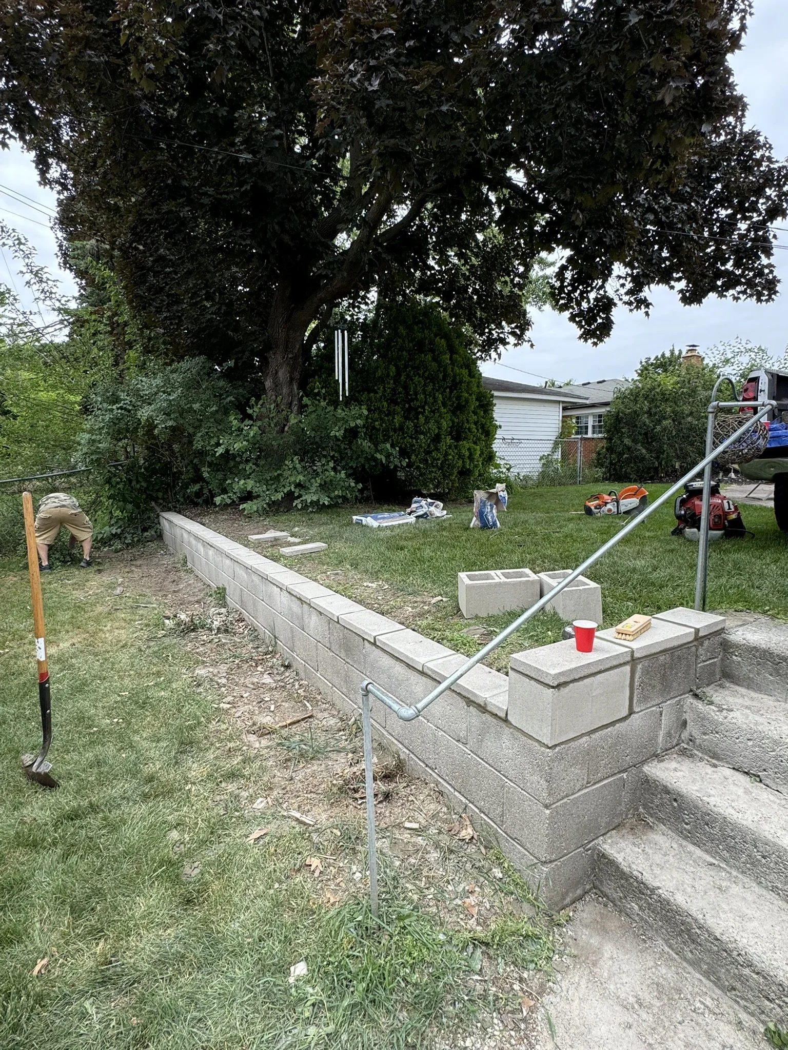 Construction site in a backyard with a worker, concrete blocks forming a retaining wall, a small set of stairs, tool bags, and gardening tools; large trees and a house in the background.