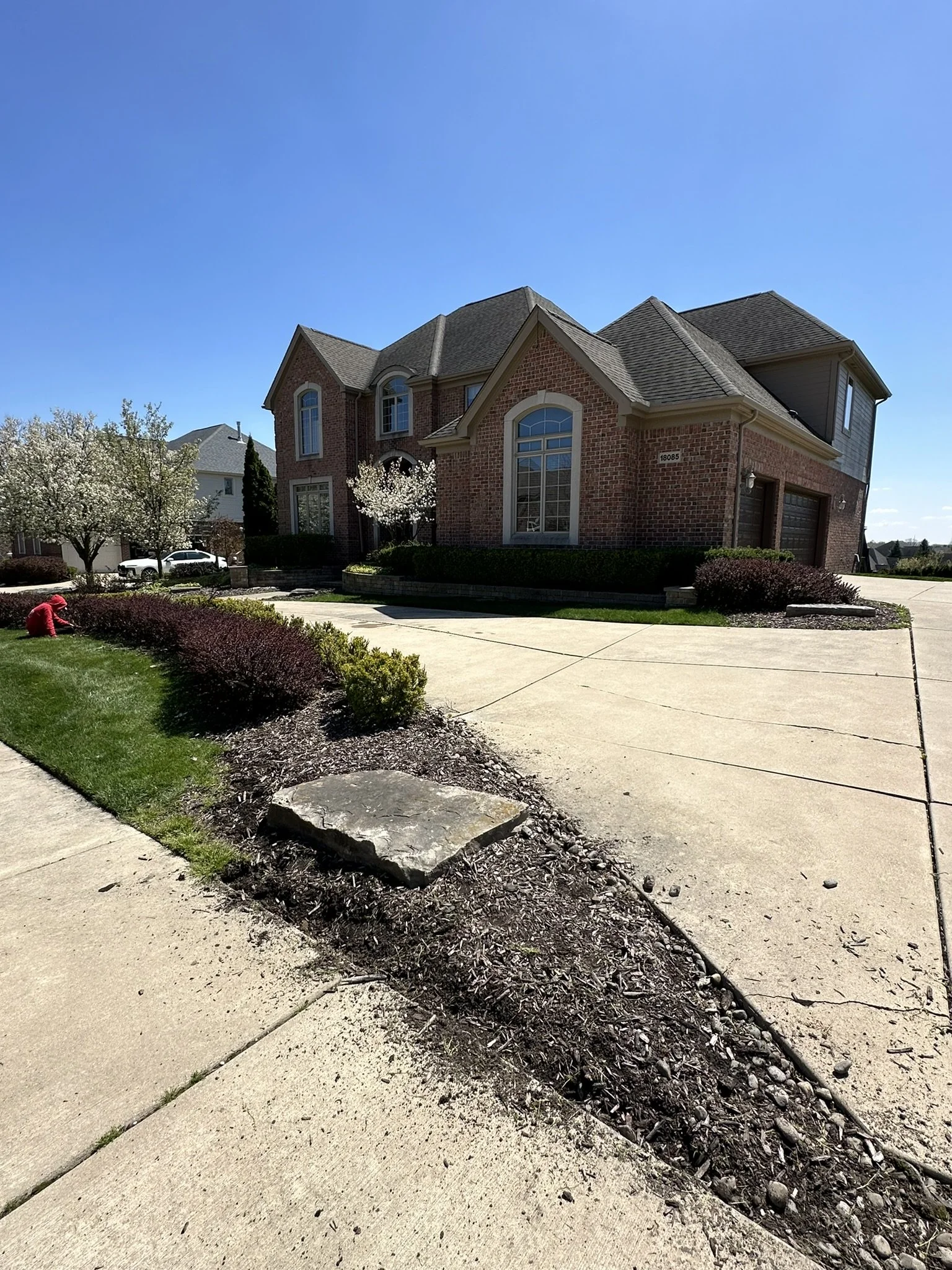 A large brick house with a well-maintained front yard, driveway, and some flowering trees in a suburban neighborhood.