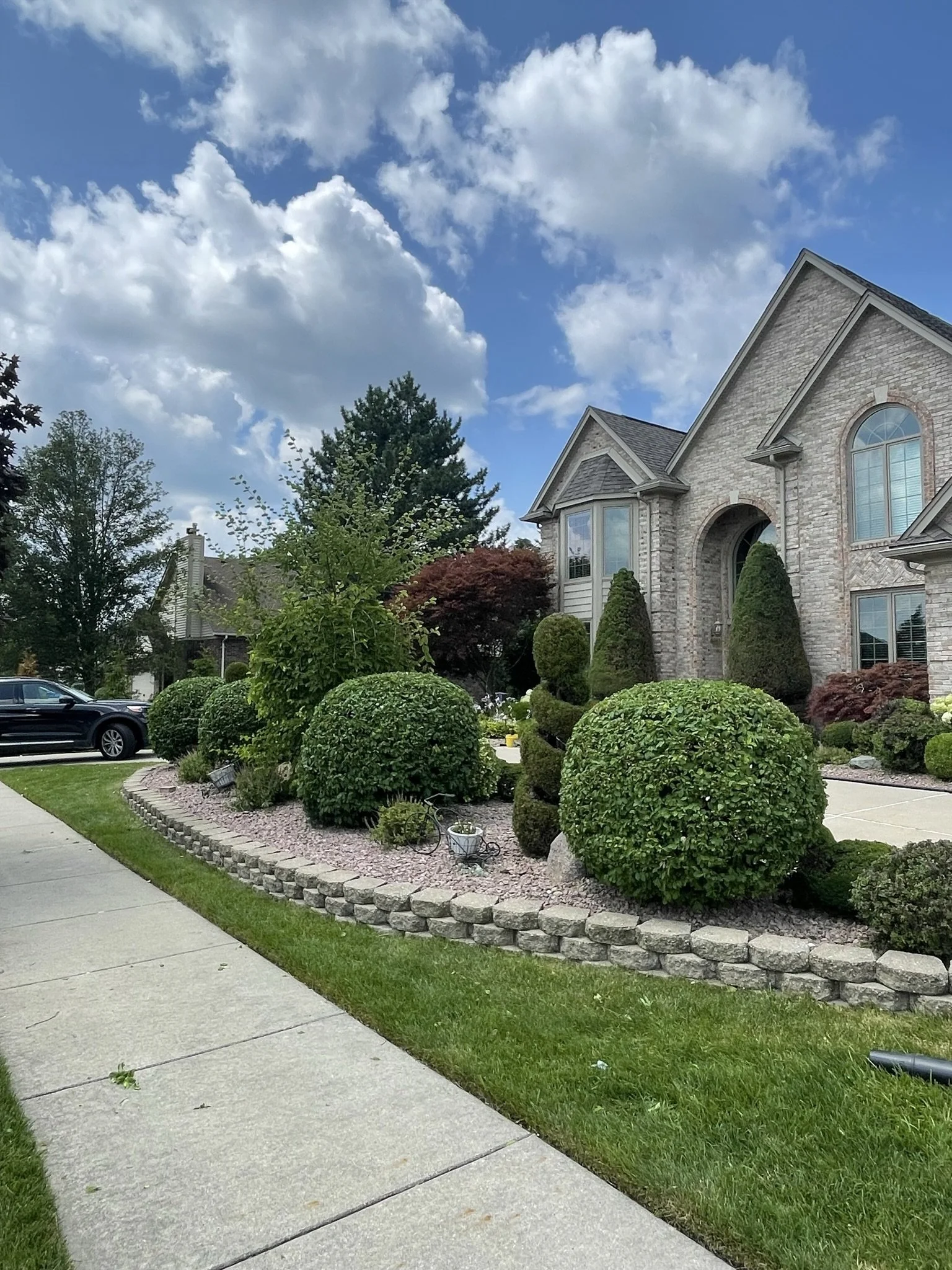 Front yard of a large house with neatly trimmed bushes, trees, and a stone border. A black car is parked on the driveway, with a sidewalk in the foreground. The sky is partly cloudy with blue sky visible.