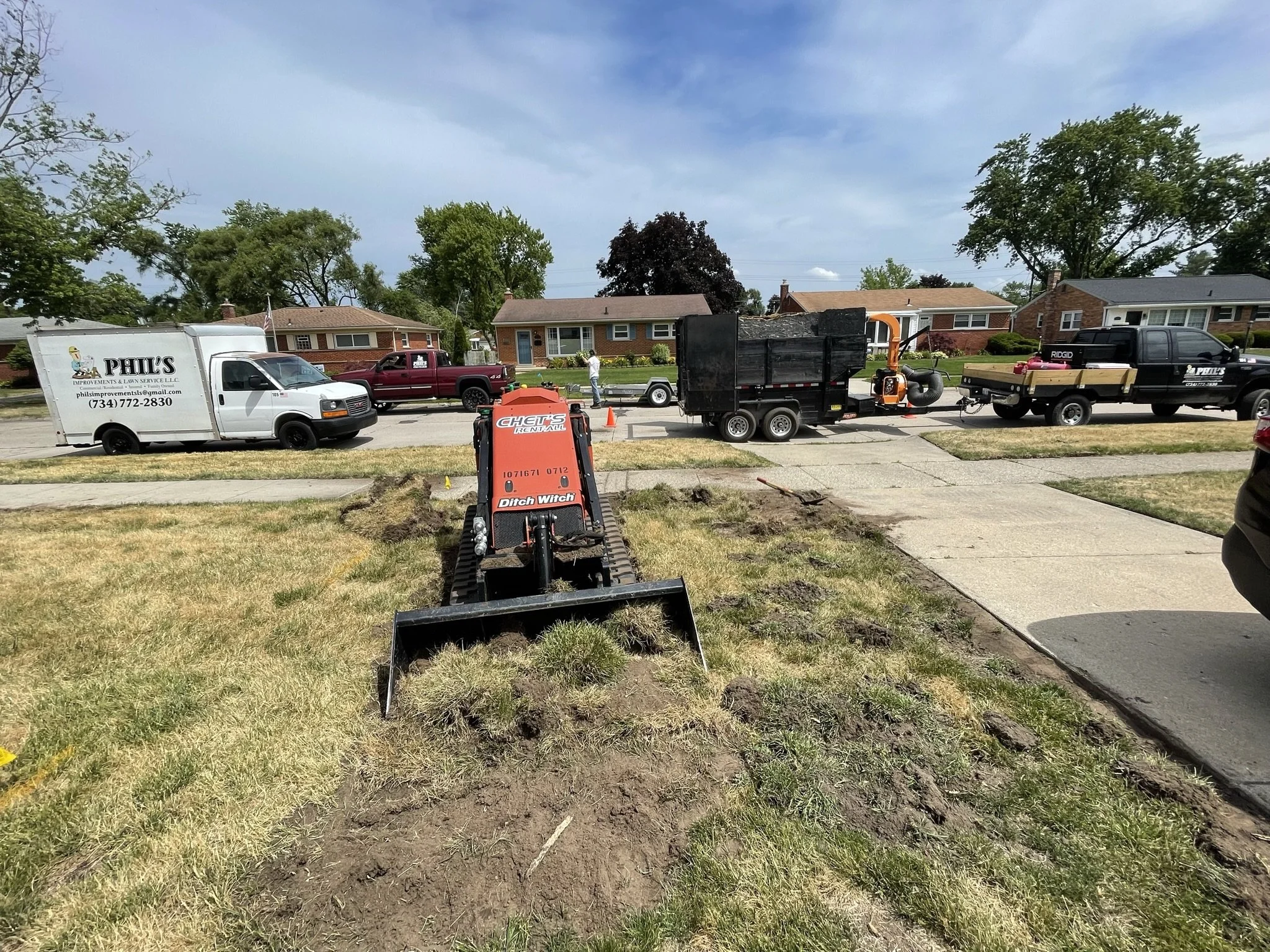 Construction crew working on sidewalk repair with equipment and multiple parked trucks on a suburban street.