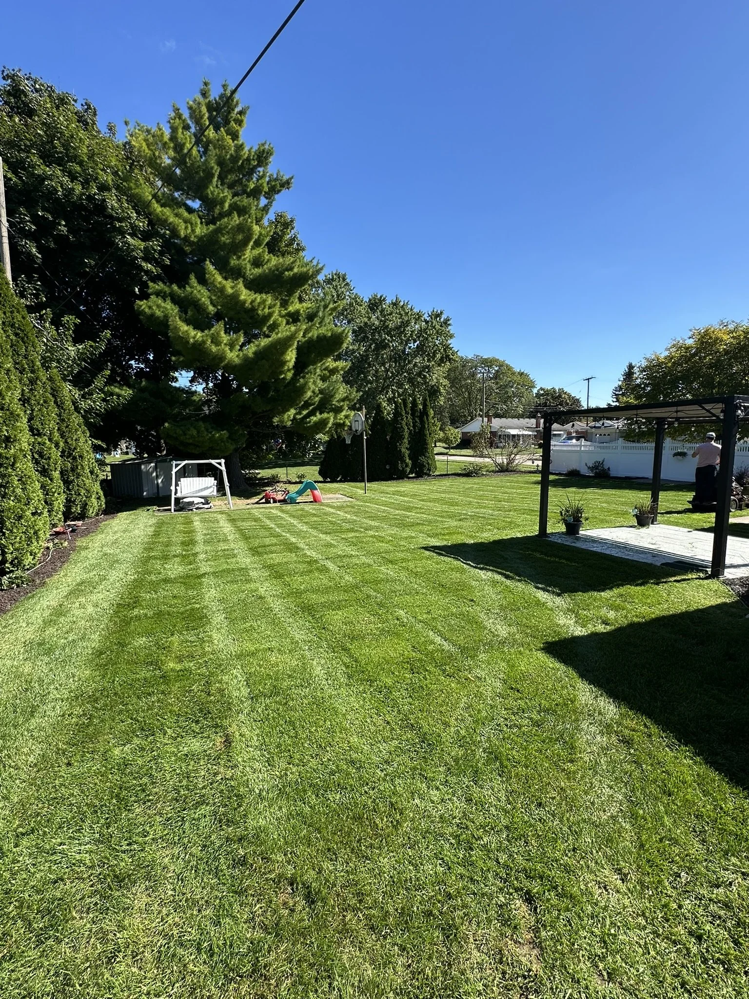 A well-maintained backyard with freshly mowed grass, a large pine tree, a small children's play area, patio with potted plants, and a person sitting on a bench against a white fence under a clear blue sky.