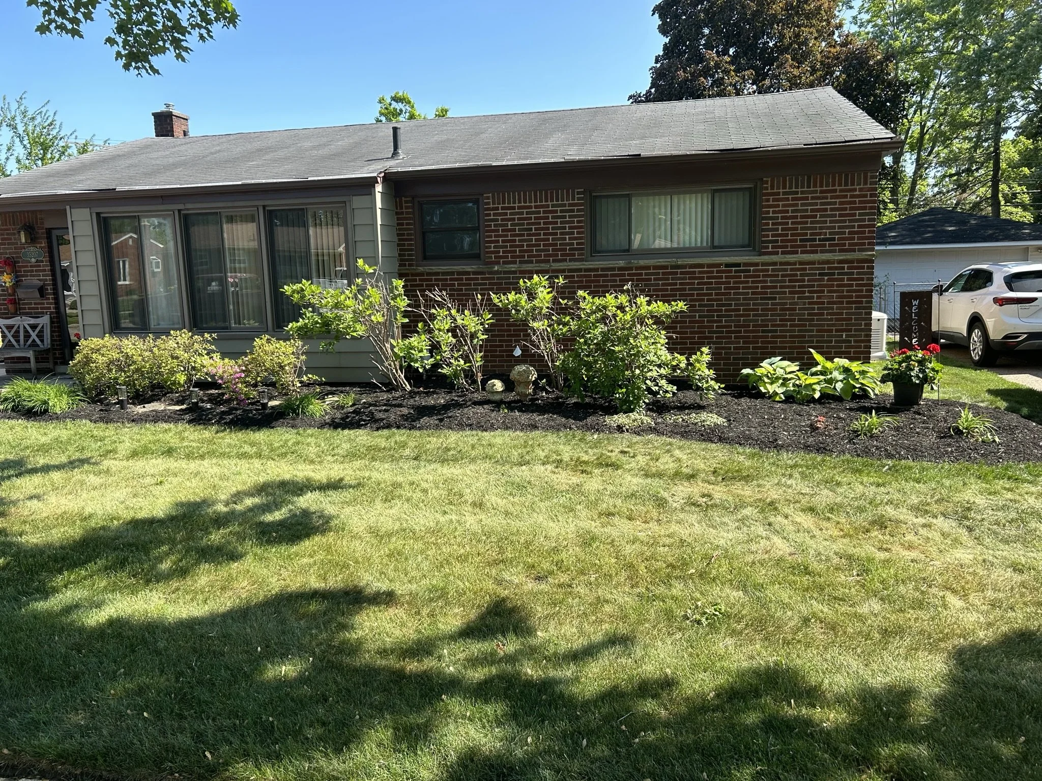 Front yard of a brick ranch-style house with a garden bed containing shrubs, flowers, and green foliage, with a lush green lawn in the foreground and a parked white SUV beside a garage in the background.