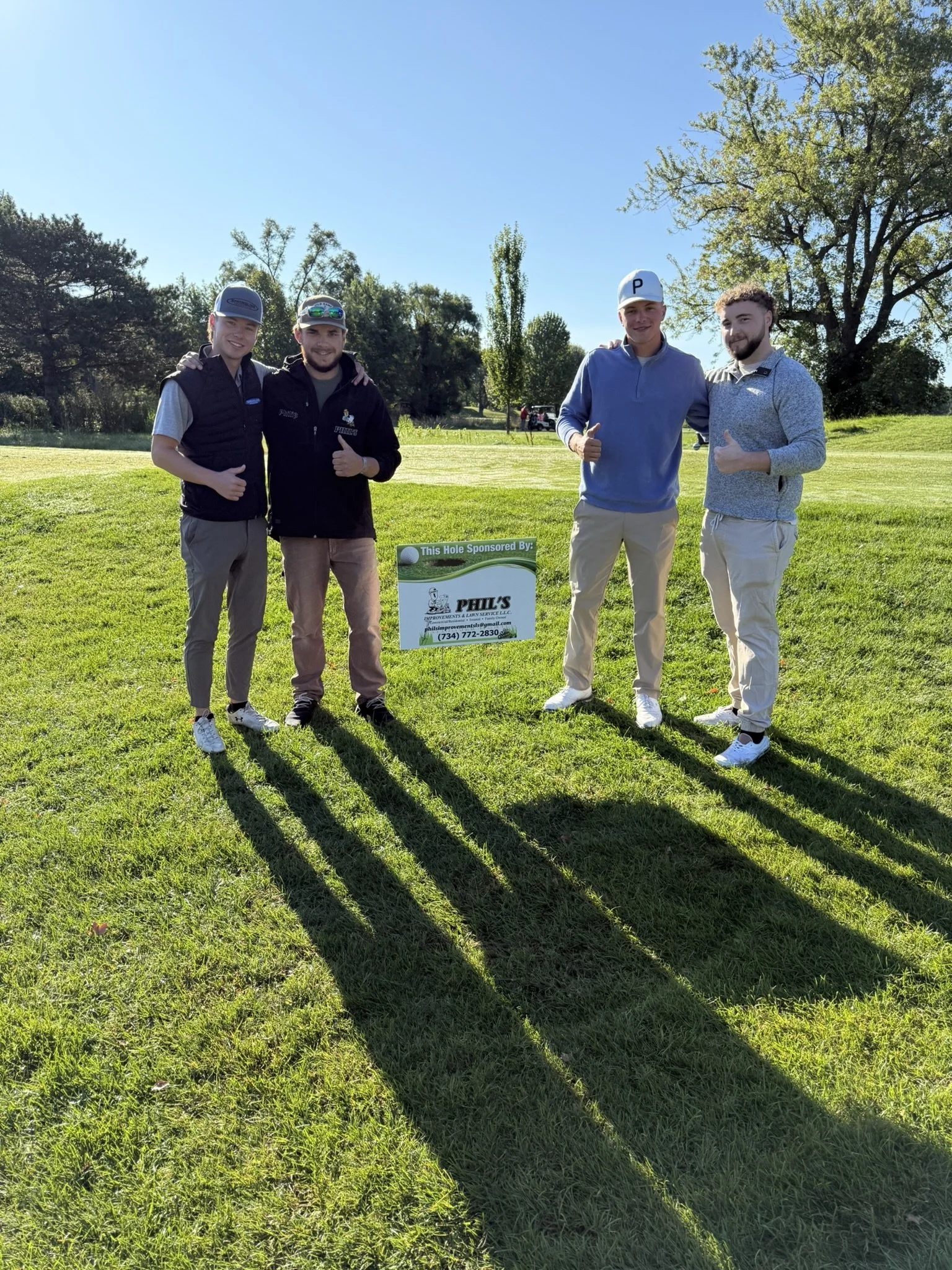 Four young men standing on a golf course beside a sign that reads, ‘This Hole Sponsored By,’ with trees in the background and long shadows on the grass.