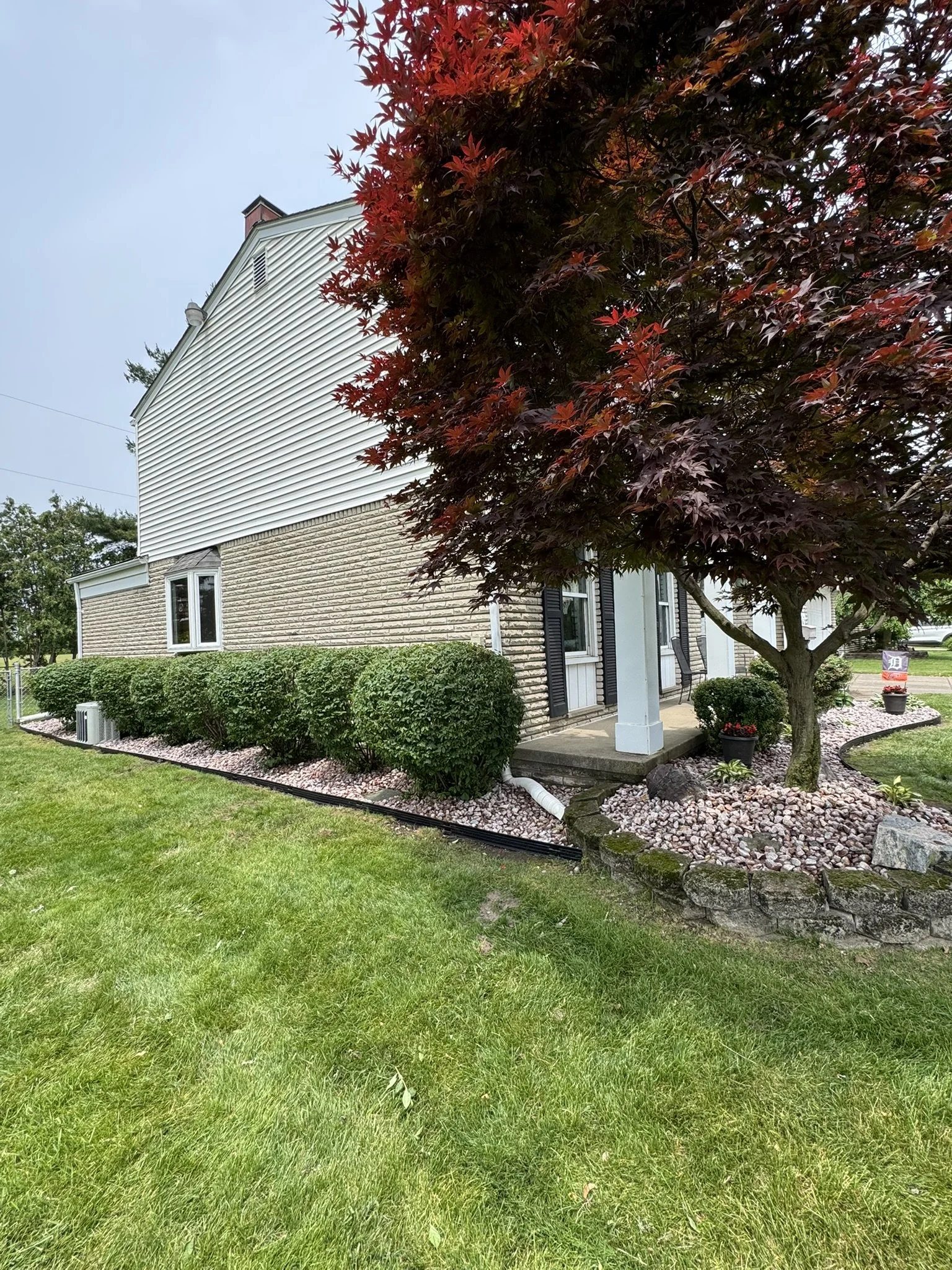 Side view of a house showing part of the lawn, a tree with red leaves, and neatly trimmed bushes with pink decorative rocks around them.