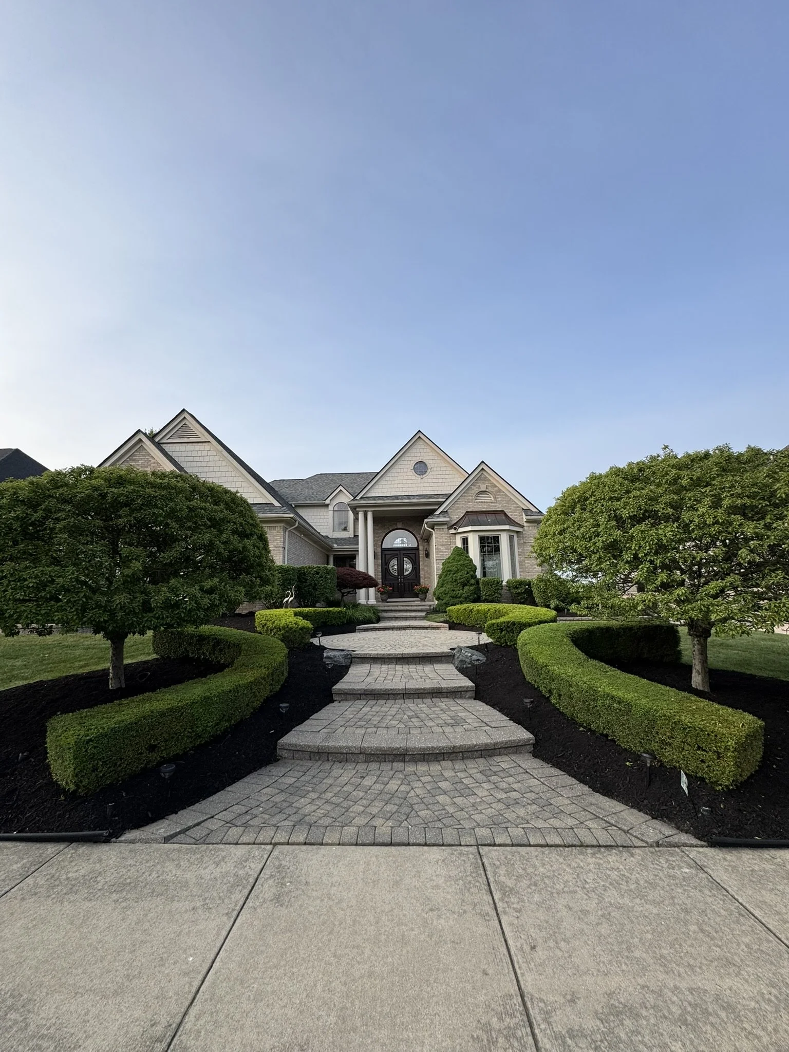 Front view of a large, elegant house with a paved pathway leading to the front door, surrounded by well-maintained trees and bushes, under a clear blue sky.