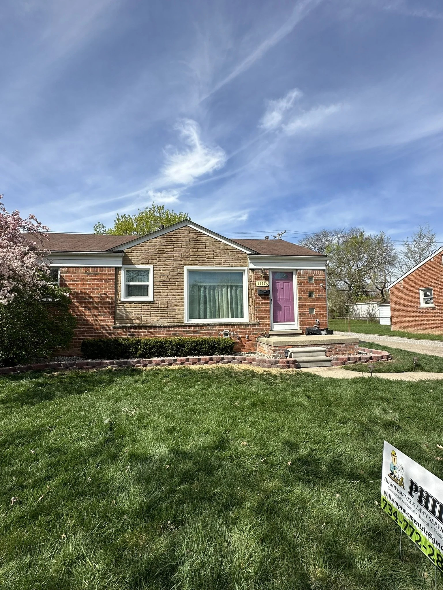 Front view of a small brick house with a purple front door, large window, and a small set of stairs leading to the entrance, surrounded by a well-maintained lawn and a 