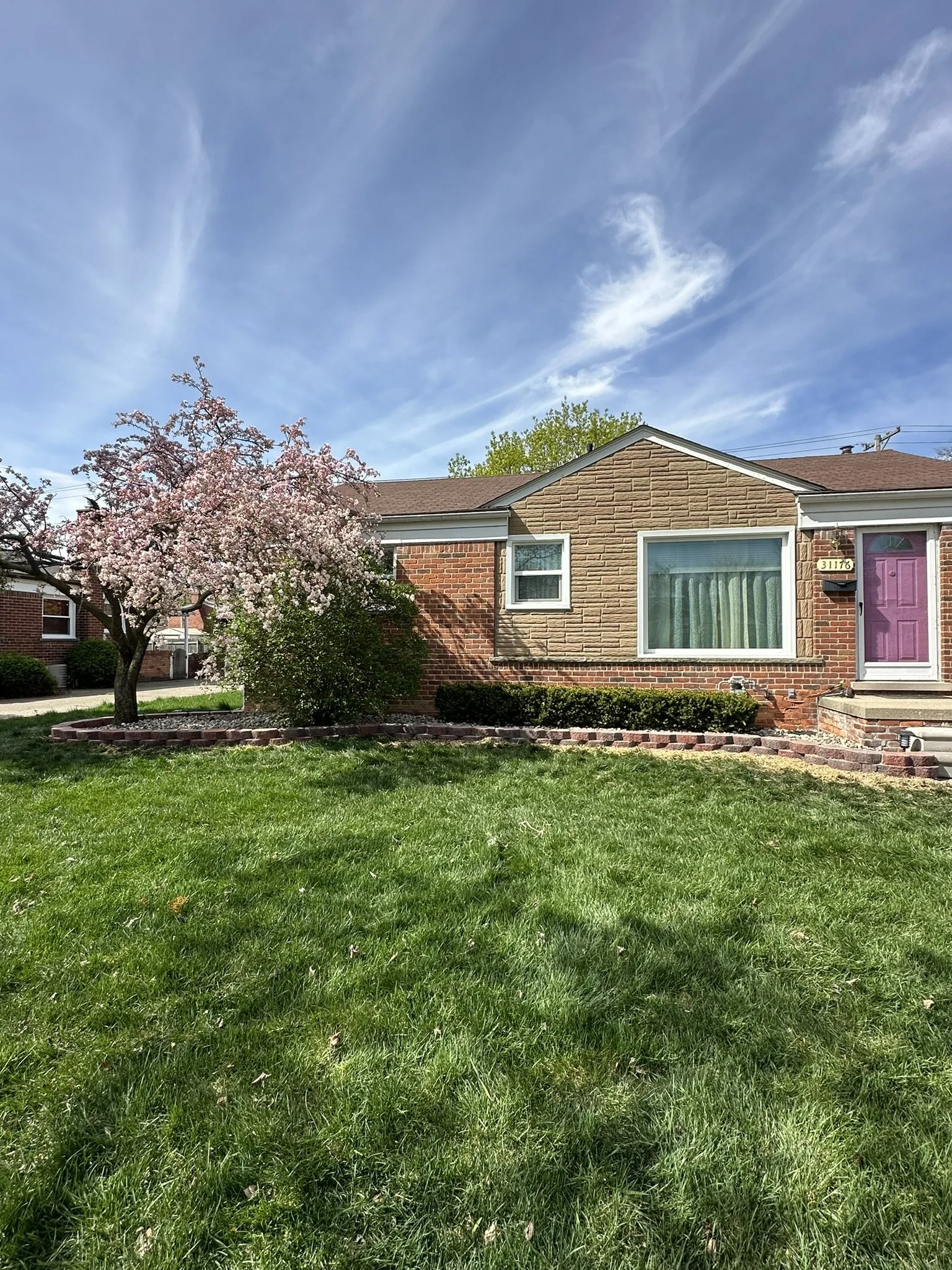 Front yard of a house with a pink flowering tree, green bushes, a well-maintained lawn, and a house with a brick and stone facade, virtual windows, and a purple front door under a clear blue sky.