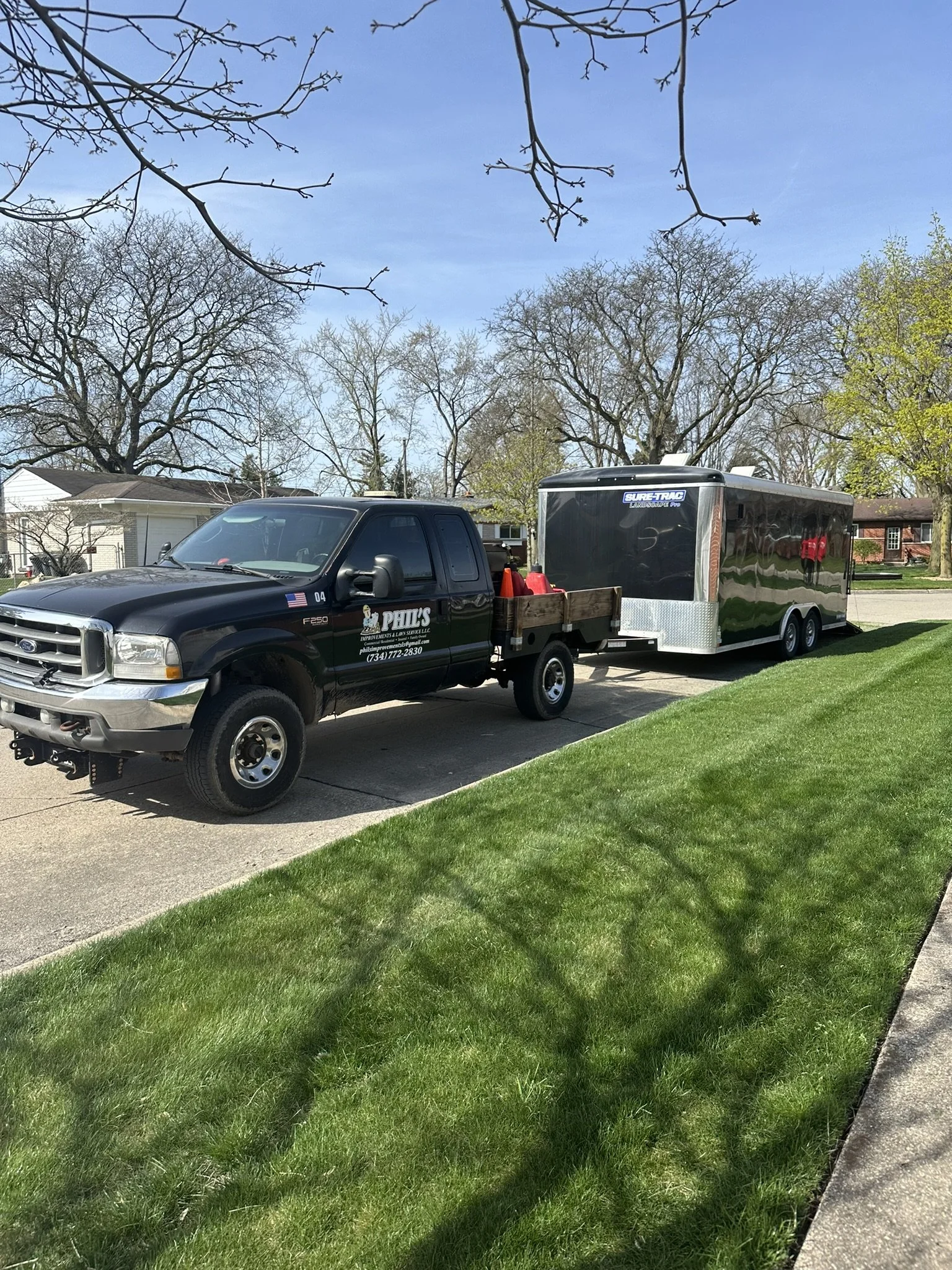 A black pickup truck with a cargo bed attached, towing a shiny black enclosed trailer in a neighborhood with green grass and leafless trees, under a blue sky.