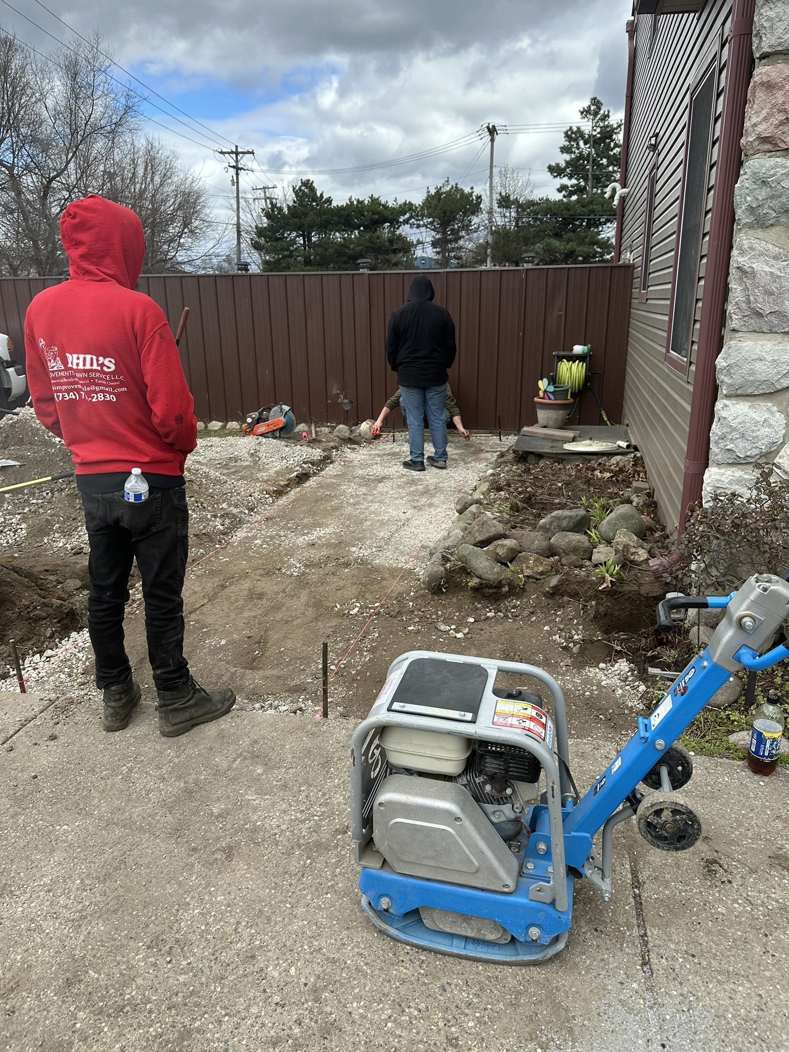 Two people working on a driveway or pathway installation, with one person in a red hoodie holding a water bottle and another in a dark hoodie and jeans working near a brown fence, tools and construction supplies scattered around.