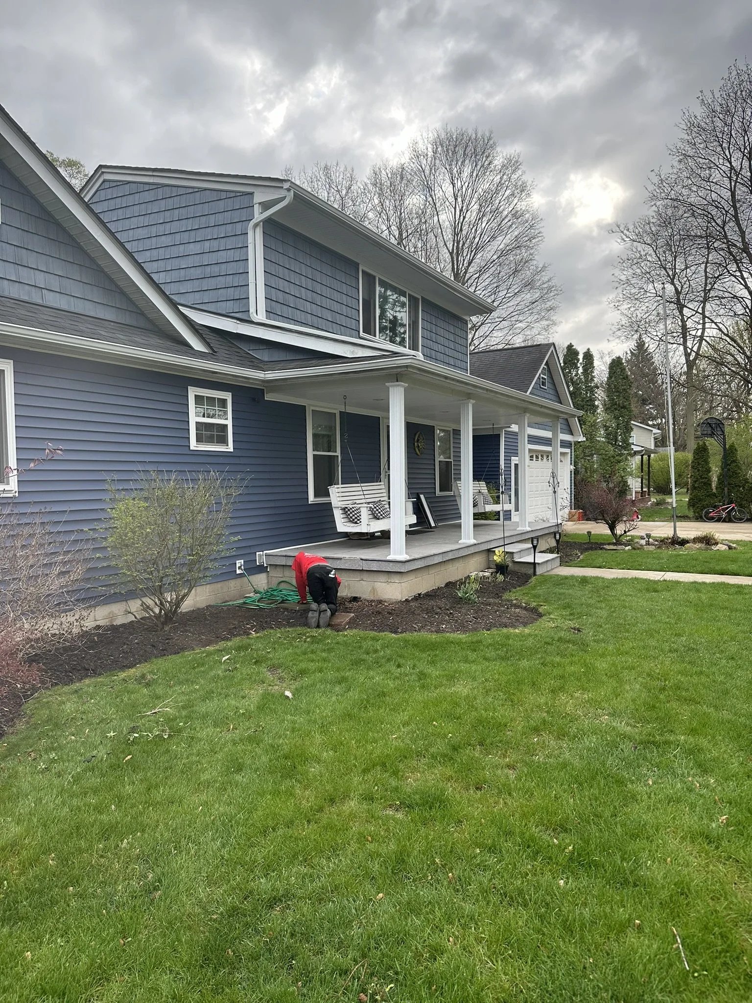 A person planting flowers or tending to a garden bed in the yard of a blue house with a porch, under an overcast sky.