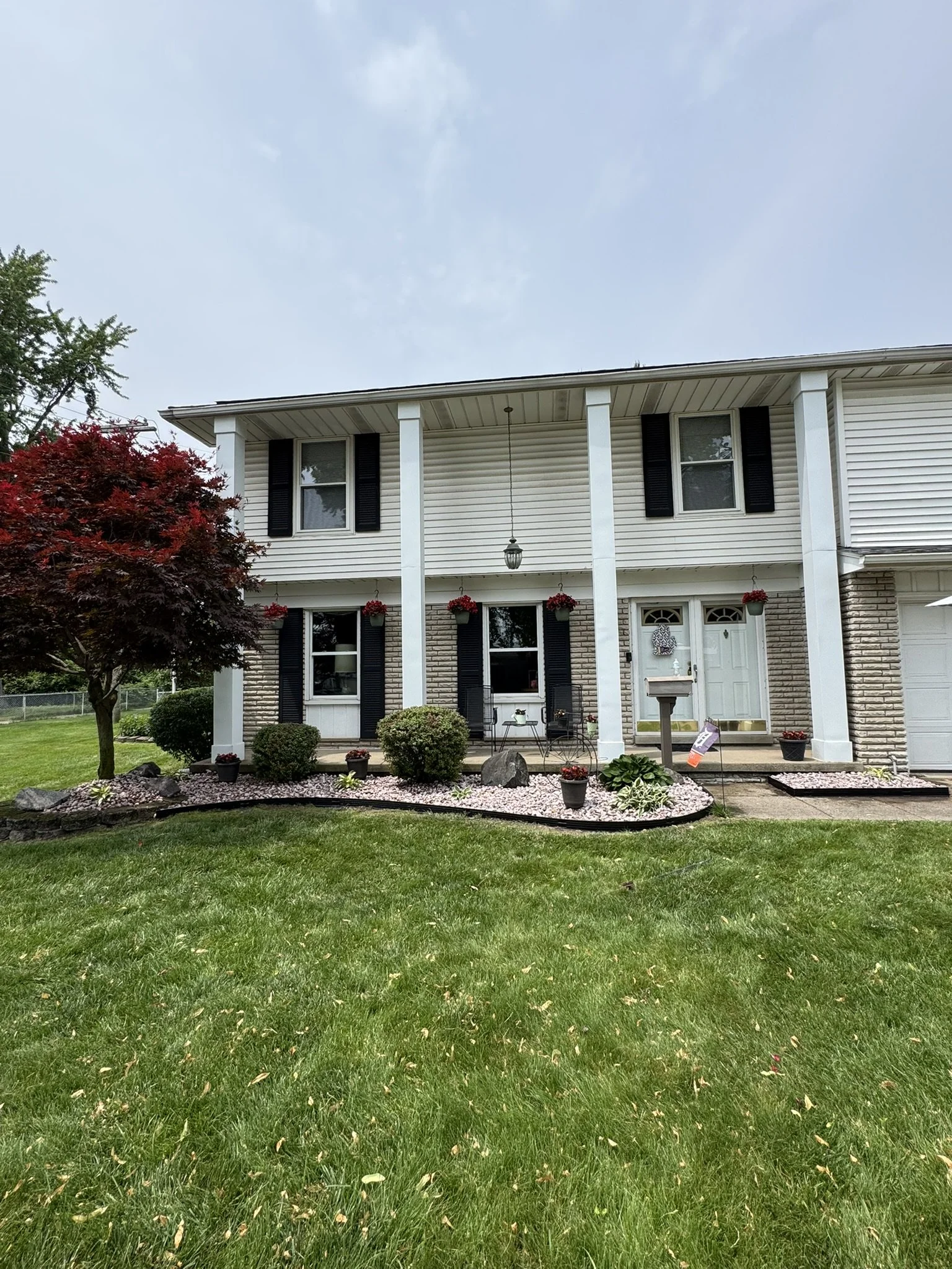 Front view of a two-story suburban house with white siding, black shutters, a front porch with potted plants, and a well-maintained front lawn with shrubs, a tree with red leaves, and decorative landscaping.