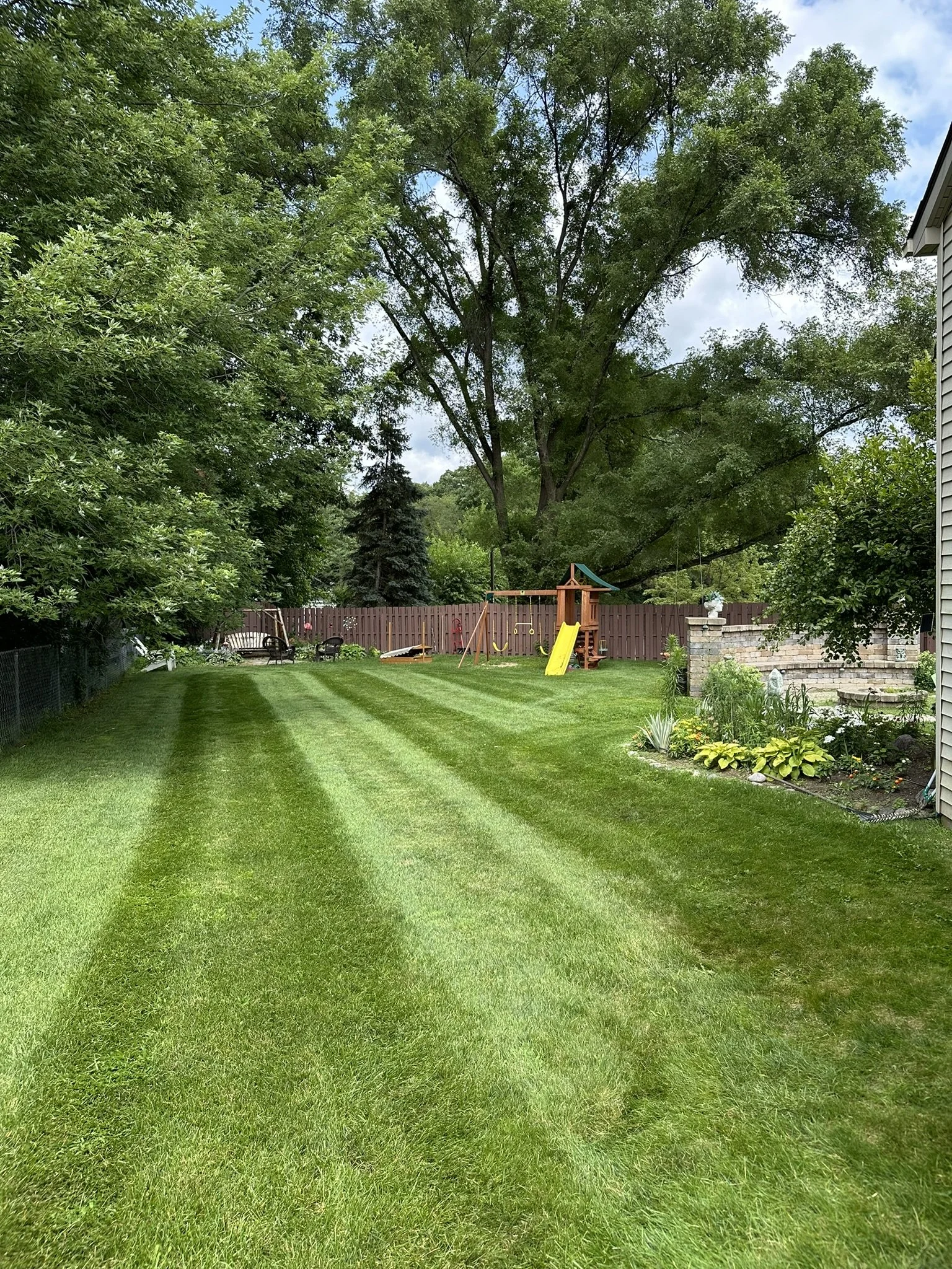 Well-maintained backyard with striped green grass, trees, a wooden playset with a slide, garden bed with plants, and a fence.
